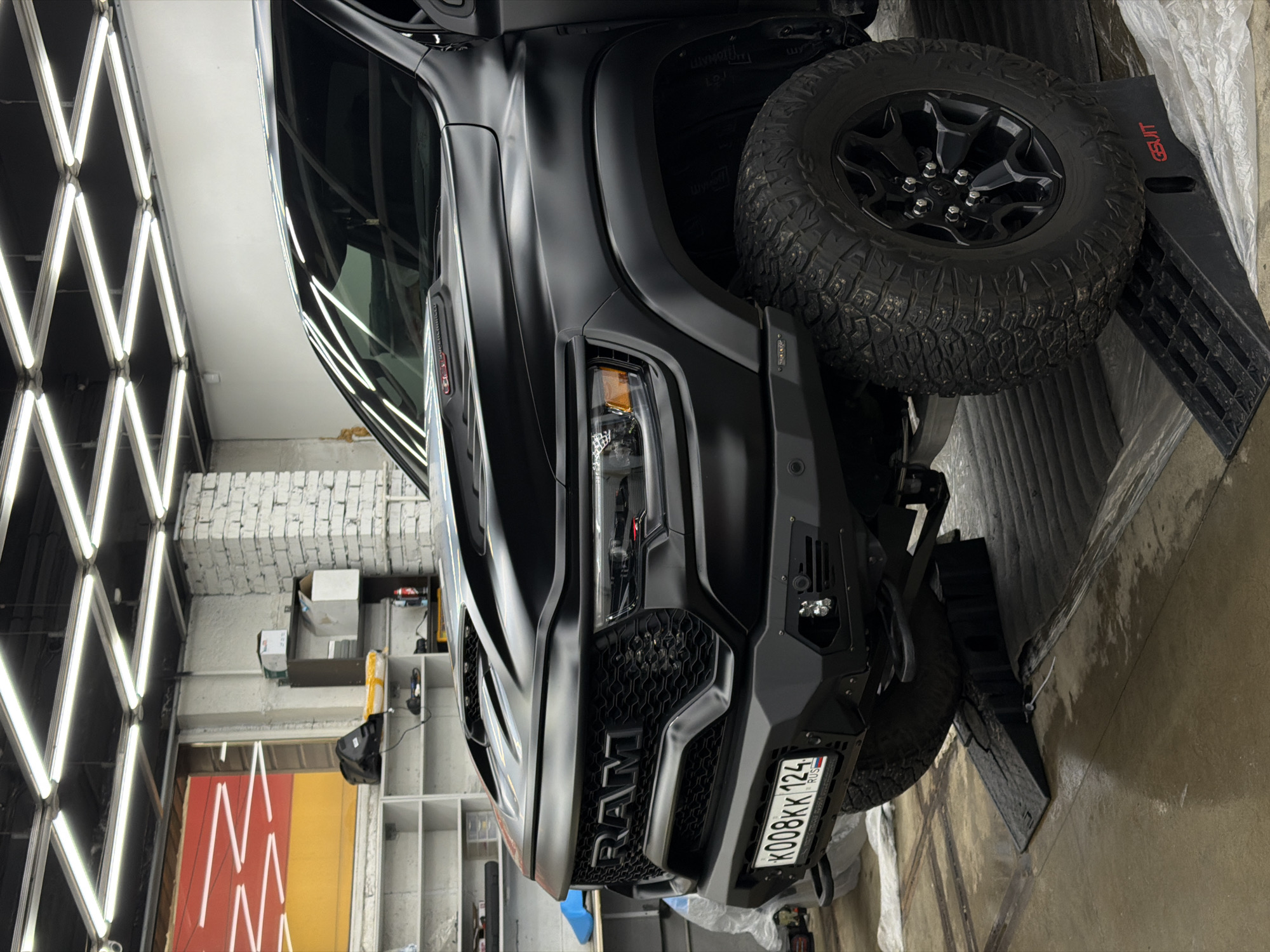 A matte black RAM truck is shown angled on a ramp within an auto service center, identifiable by fluorescent lighting and shelving. The vehicle's Russian license plate reads "K008KK 124 RUS", indicating registration in the Krasnoyarsk region of Russia. The truck is positioned with its front end elevated on a black ramp, and a tire is prominently displayed. The garage interior is visible, including shelves, tools, and colored panels.