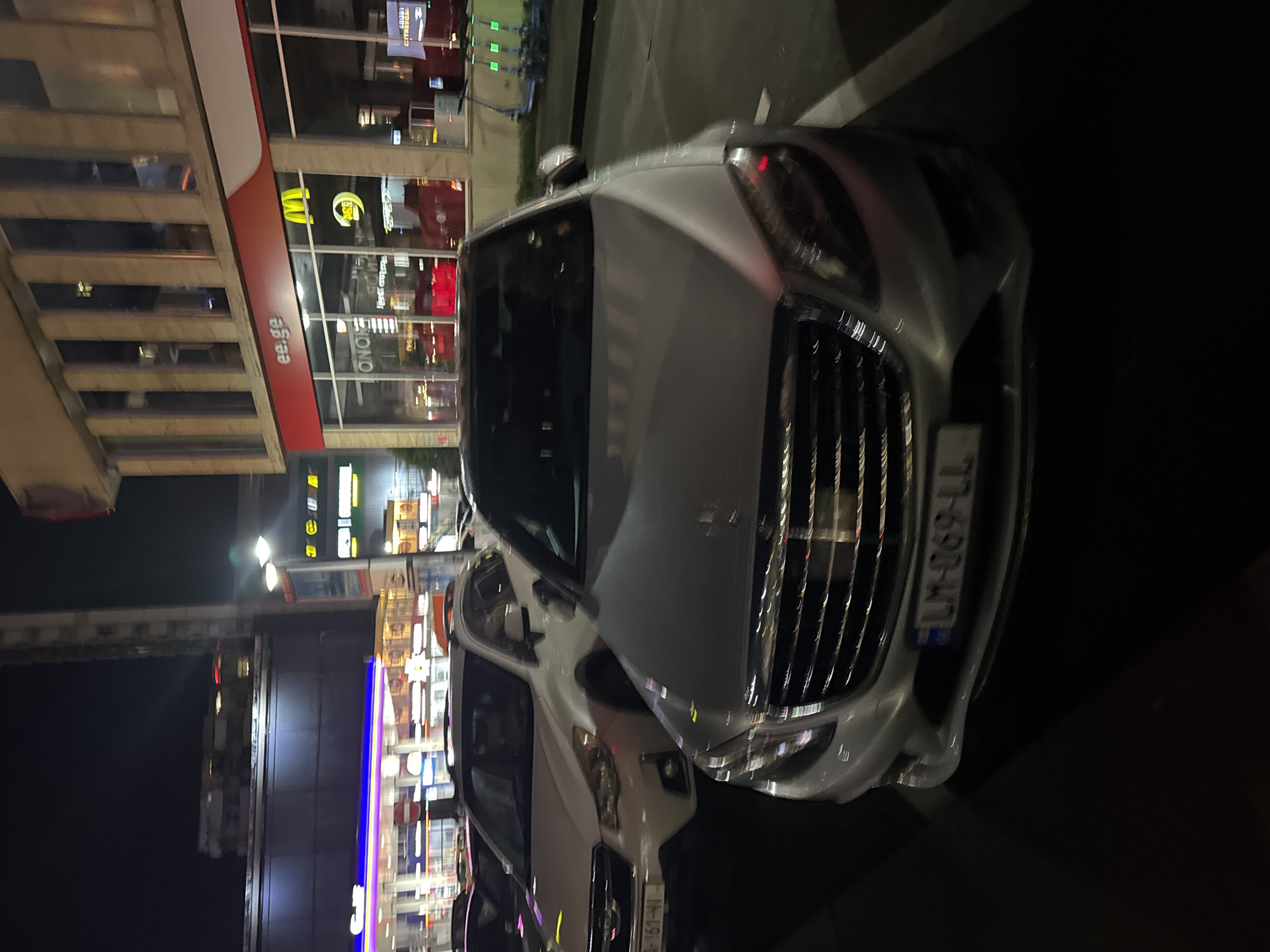 Here is a detailed description of the image:
The image presents a nighttime urban scene, focusing on the front of a silver car parked on what appears to be a city street. The car is positioned at an angle, showing its front grille, headlights, and license plate which reads "LM-069-LL". The background includes buildings with brightly lit storefronts, displaying signs for various businesses. The architecture seems modern, with glass facades and visible internal structures. The lighting suggests an urban environment with artificial illumination. The street is dark and the lighting conditions are low, creating a stark contrast between the foreground and the background. The overall impression is one of a typical urban setting at night, with the car as the central subject of the scene.