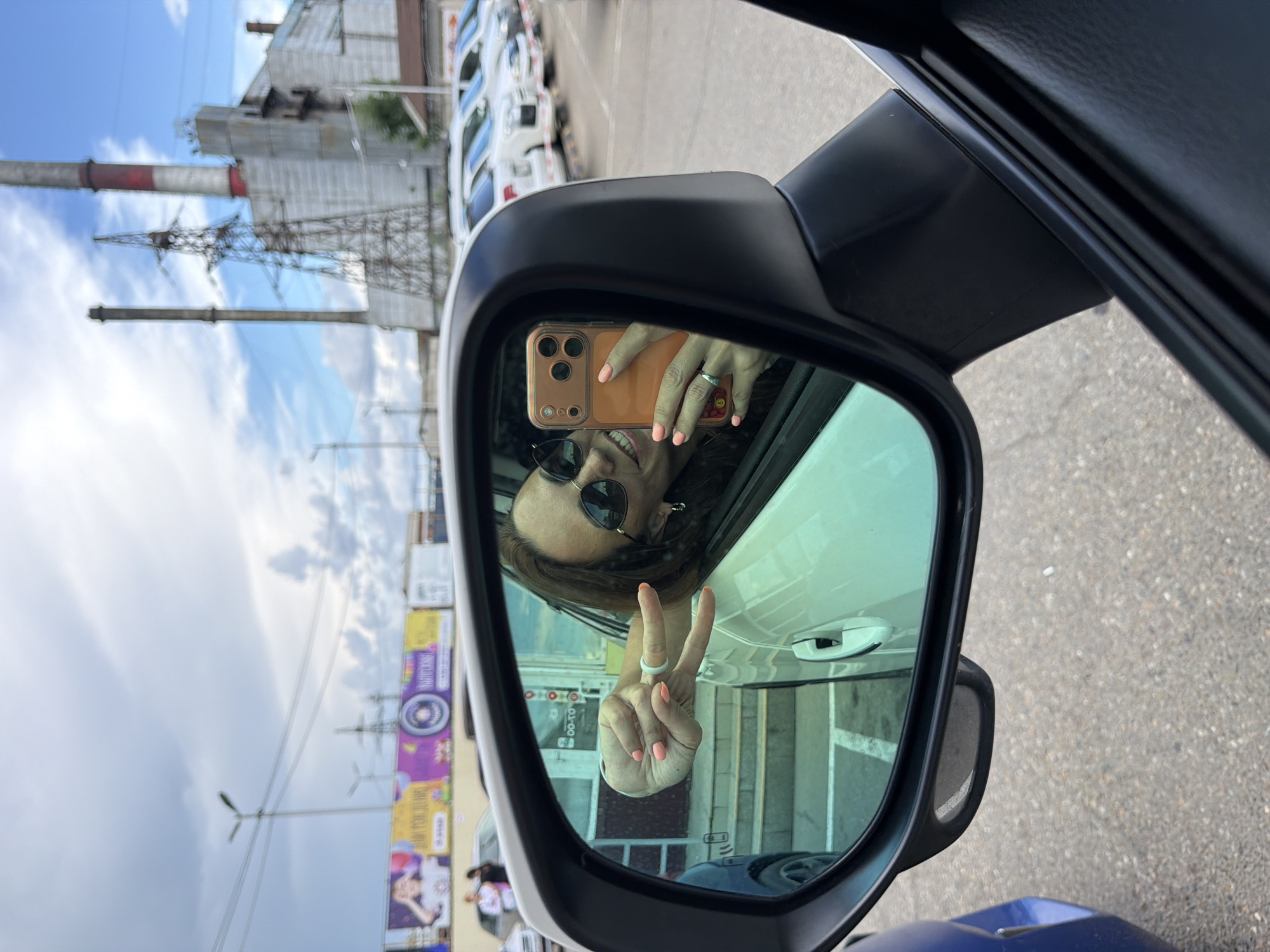 A woman is reflected in a car's side mirror, taking a selfie with her smartphone. She is wearing sunglasses and making a peace sign with her left hand, which has bright coral nail polish. A ring is visible on her ring finger. The background visible in the mirror shows a parked car, a building with stairs, and a billboard advertising a children's party. Beyond that, industrial structures including smokestacks and power lines are visible against a cloudy sky. The scene is set in Birobidzhan, Russia.