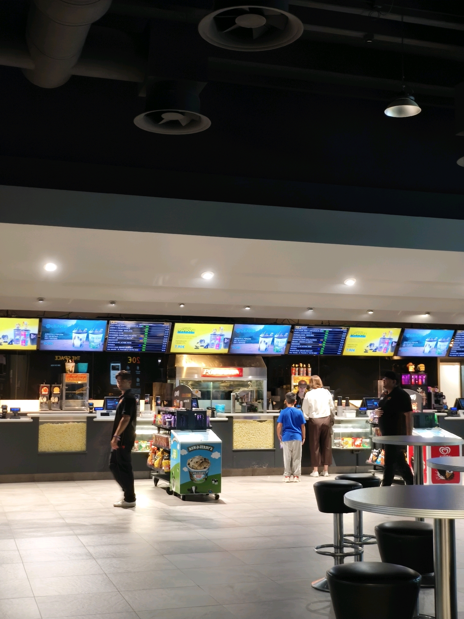 The image depicts the concession stand area of a cinema, likely located in the city of Chiugiana, Italy, based on the information provided. In the foreground, several round tables with black stools are arranged on a tiled floor. Behind these tables, a long counter serves as the concession area.
Several individuals are visible at the counter. A young person in a blue t-shirt and grey pants stands facing the counter, possibly ordering. To their right, a woman in a white top and brown pants is also at the counter, interacting with an employee. Another employee, wearing a dark uniform, is visible to the right of this group, appearing to be serving customers. A lone individual in a dark t-shirt and black pants stands further down the counter on the left side of the image, looking towards the concession area.
Above the counter, large digital screens display menus and movie-related advertisements. One screen prominently features "Minions" imagery. Various concession items, including popcorn containers, are visible on the counter and behind it. A refrigerated display case with ice cream, likely Ben & Jerry's, is also present. The ceiling features modern lighting and ventilation systems. The overall environment suggests a brightly lit, modern cinema lobby.