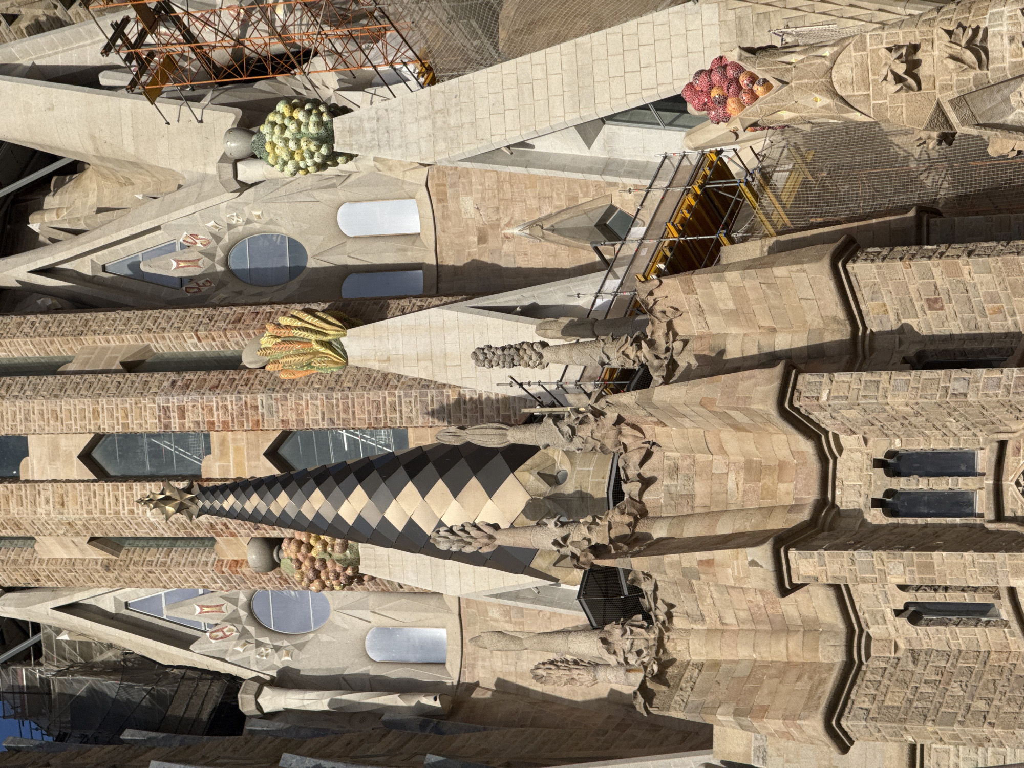 The image displays a detailed close-up of the Sagrada Familia basilica in Barcelona, Spain, focusing on its intricate stone architecture and decorative elements. The facade features pointed arches, diamond-patterned spires, and sculpted fruit-like motifs, possibly representing grapes or pomegranates, in vibrant colors of red, orange, and yellow. Tall, narrow windows with dark panes are visible, interspersed with stone carvings and textured brickwork. Scaffolding and netting suggest ongoing construction or maintenance. The lighting indicates it is daytime, with strong shadows cast by the architectural elements, highlighting the three-dimensional nature of the facade. No people are visible in the frame.