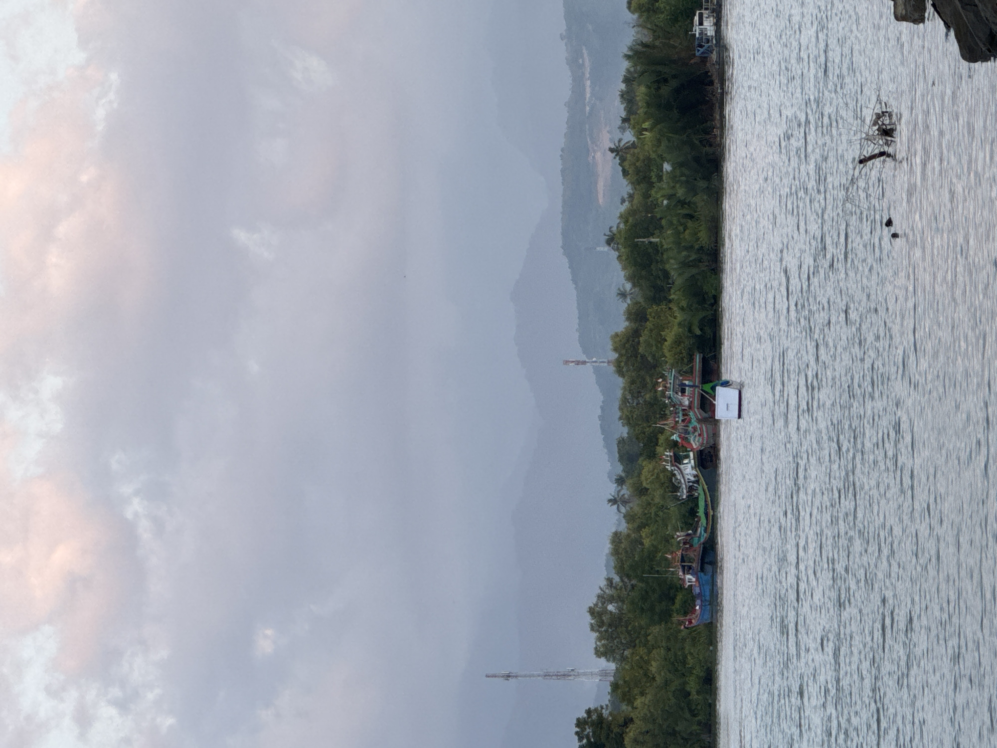 The image shows a body of water in front of a lush green coastline with several boats moored. In the background, a range of misty mountains can be seen under a cloudy sky. On the left side of the coastline, a tall communication tower stands prominently. The water is calm with gentle ripples, and a small white buoy floats in the middle ground. The overall scene depicts a tranquil coastal landscape.