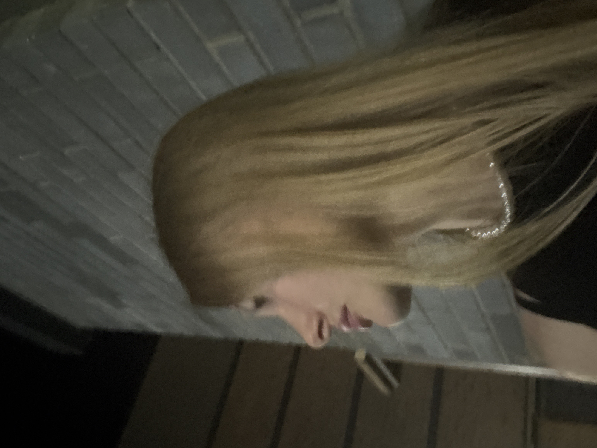 The image shows a young woman with long blonde hair, wearing a black top and a pearl necklace. She is looking to her left, with her profile visible. The background is a brick wall and a darker area which appears to be a building. The lighting suggests it is nighttime or a dimly lit indoor space. The context does not provide any information to determine the location in the city of Perm, Russia.