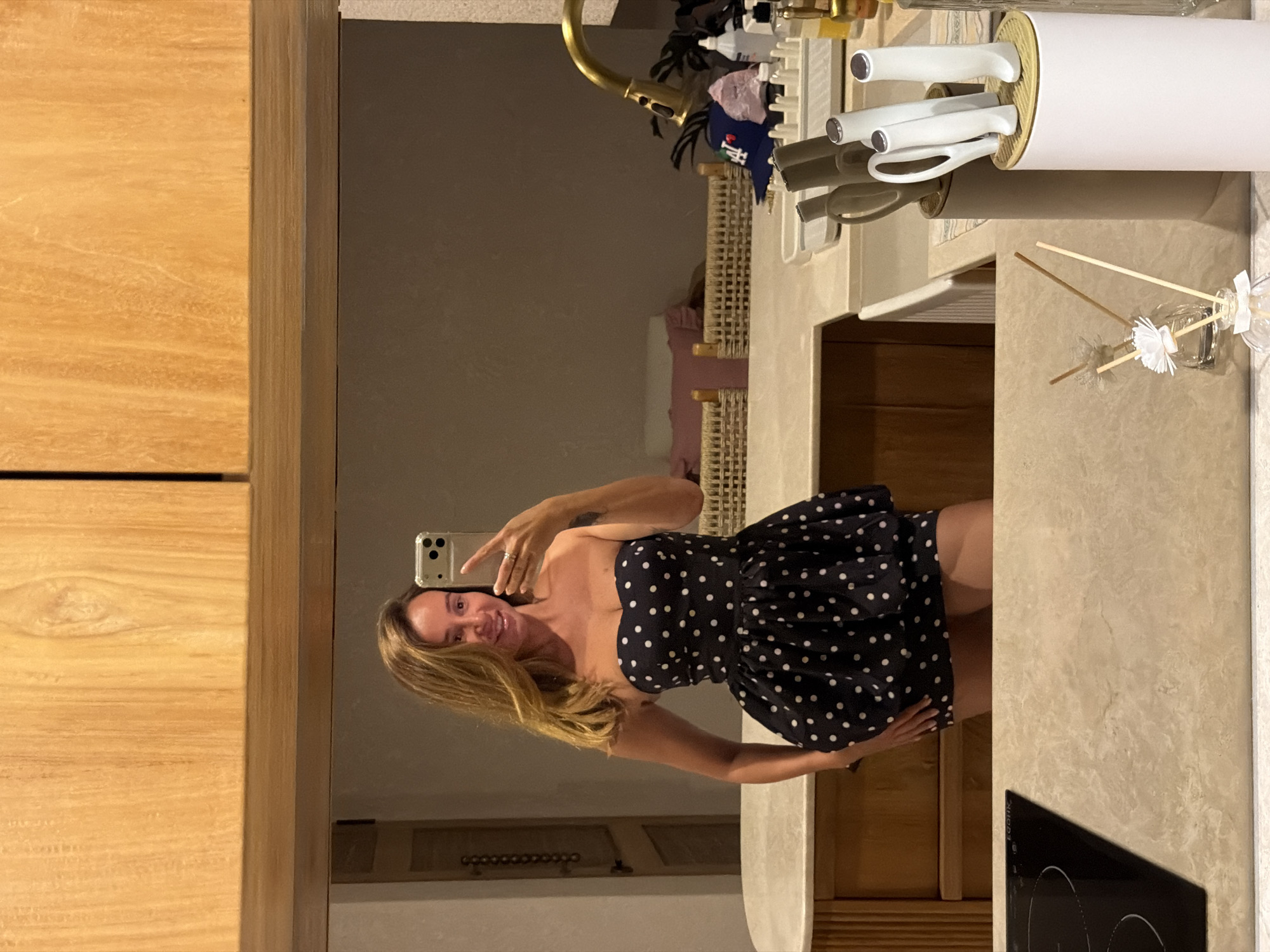 A woman is taking a mirror selfie in what appears to be a modern kitchen. She is wearing a black strapless dress with white polka dots, which has a sweetheart neckline and flares out at the waist. The dress is short, ending above her knees. She is holding a silver smartphone in her right hand to take the picture. Her left hand is placed on her hip.
The background shows a kitchen counter with a sink, a faucet, a dish drying rack with some items on it, and a knife block with several knives. There are also bottles of cleaning supplies or toiletries on the counter. Above the counter, there are wooden cabinets. A stovetop is visible in the foreground on the left. The overall environment suggests a residential kitchen. The reflection in the mirror also reveals a doorway or a closet door on the left side of the frame. There is no indication of the location being in the city of Mengwi, Indonesia.