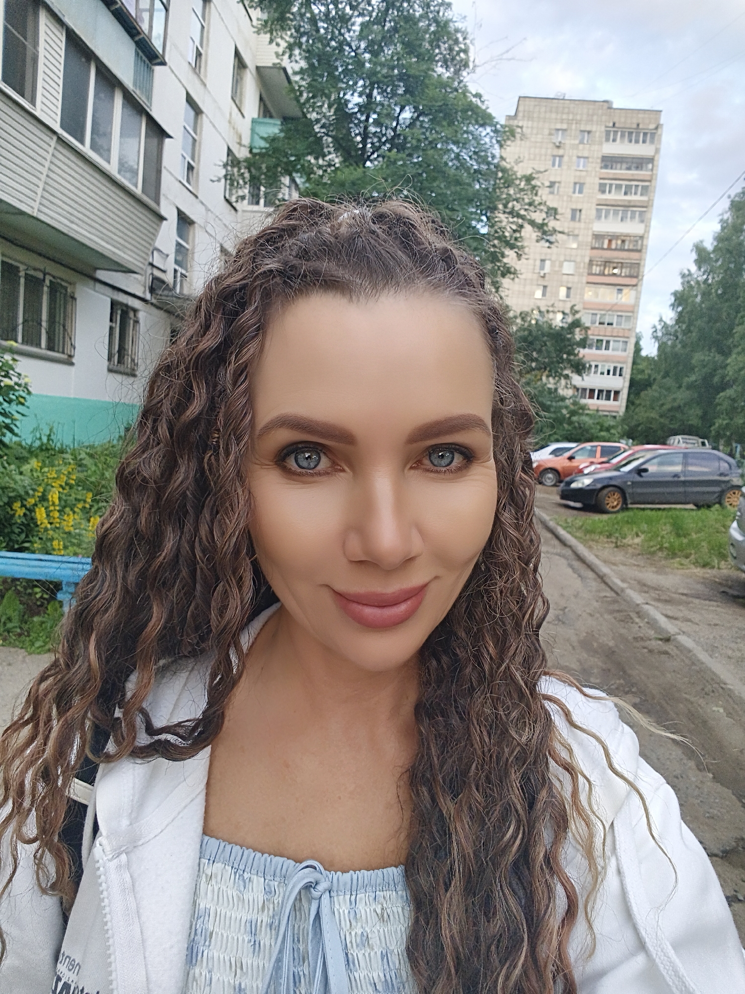 A medium shot selfie features a woman with curly brown hair and blue eyes, looking at the camera. She is wearing a white zippered jacket over a light blue smocked top with a tie closure. Her makeup includes eyeshadow and lipstick. The background shows a residential area in Chaikovsky, Russia, with a light-colored apartment building on the left and a taller, beige apartment building on the right. Trees and parked cars are visible between the buildings and along a paved road. The sky is overcast.