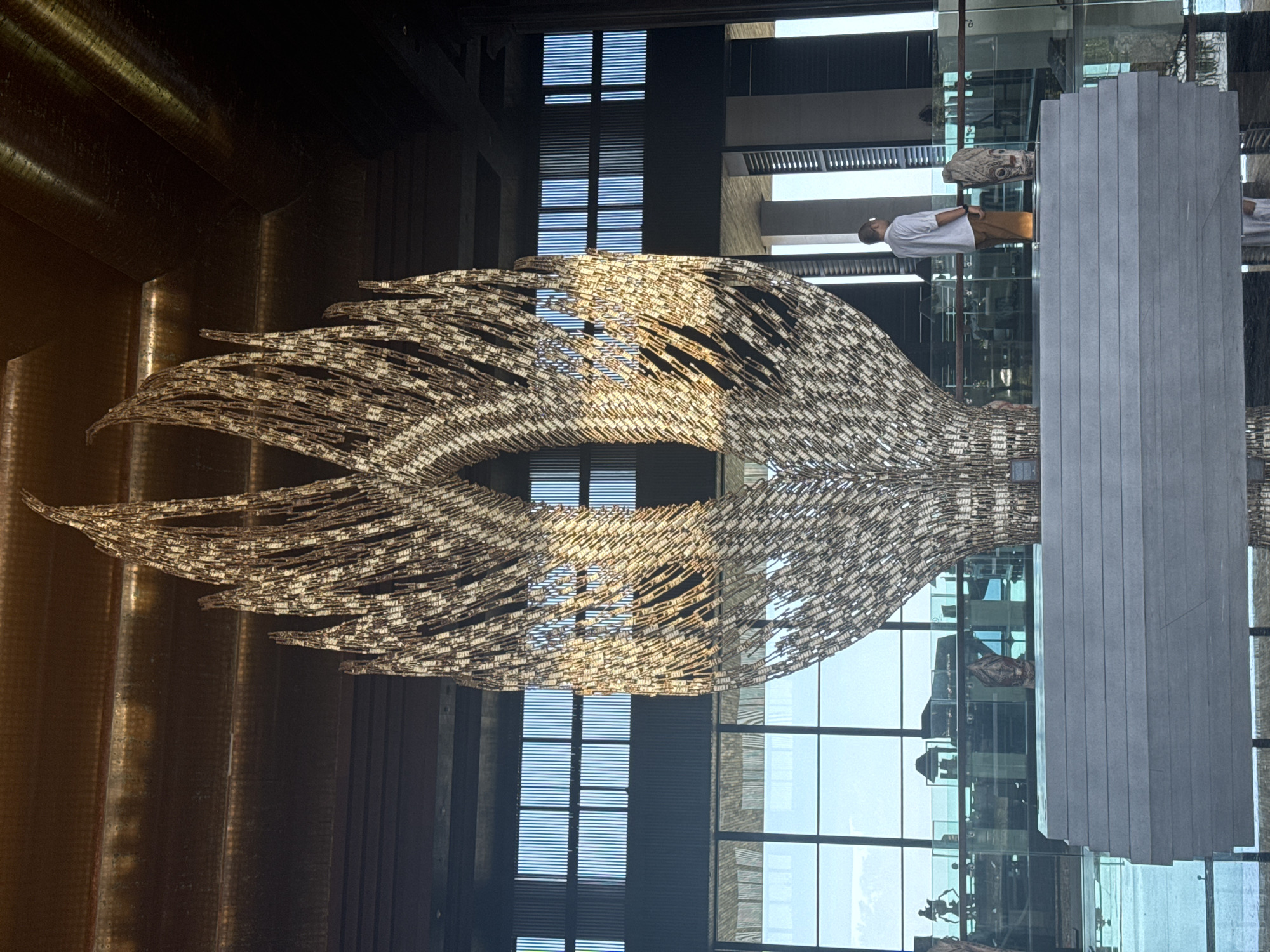 The image shows a large, intricate chandelier or light installation, made of what appears to be small, reflective pieces, resembling feathers or scales. The installation is symmetrical, wing-like, and dominates the foreground.
Behind the installation, there are architectural elements suggesting a modern building interior. A large glass wall with visible mullions allows a view of the outside, potentially indicating a lobby or atrium area. There are linear, dark structures that might be window blinds or architectural features, adding depth to the background.
In the reflection of the glass wall, figures of people are visible. One man is wearing a white shirt and tan pants. There are also architectural details reflected, suggesting the building continues above and below the camera's viewpoint.
The location is the Renaissance Bali Uluwatu Resort & Spa in Kuta Selatan, Indonesia.