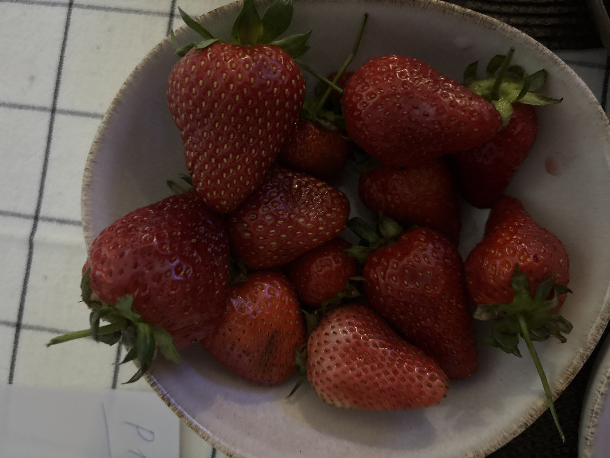 A close-up, overhead shot depicts a bowl filled with strawberries. The bowl is light in color with a textured edge. The strawberries are various shades of red, some with green stems and leaves still attached. They appear fresh and ripe. The bowl sits on a surface that is partially covered by a checkered white and grey fabric. Part of a white object with the handwritten letter "P" is visible in the bottom left corner. The overall scene suggests a domestic or culinary setting. The location in Saint Petersburg, Russia is unknown.