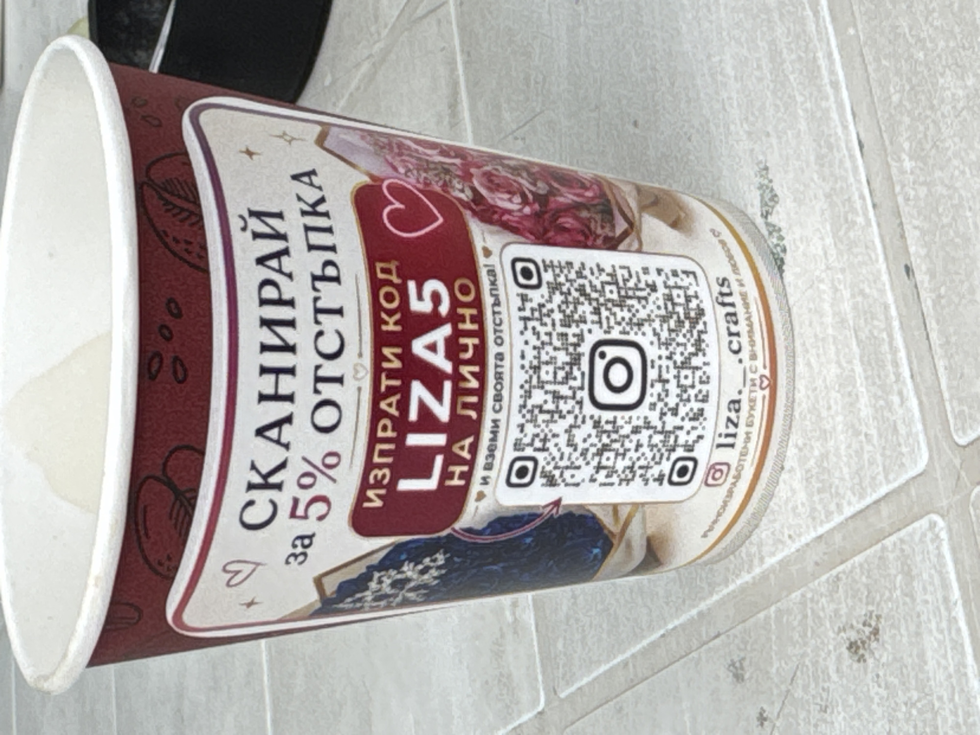 The image displays a close-up of a disposable paper cup, likely from a coffee shop, situated on a tiled surface. The cup is decorated with a predominantly red and white design featuring floral imagery and text in Bulgarian. A prominent feature is a QR code located centrally on the cup. Above the QR code, the text "ИЗПРАТИ КОД LIZA5 НА ЛИЧНО" is visible, and below it, "И ВЗЕМИ СВОЯТА ОТСТЪПКА!". The cup also has a white rim. The tiled surface appears to be an outdoor setting, possibly a patio or sidewalk, with visible grout lines and some light debris. The background is blurred, but suggests an urban or outdoor environment. The overall impression is that this is a promotional item from a business. There is no specific indication of the city of Sredets, Bulgaria within the visual information provided.