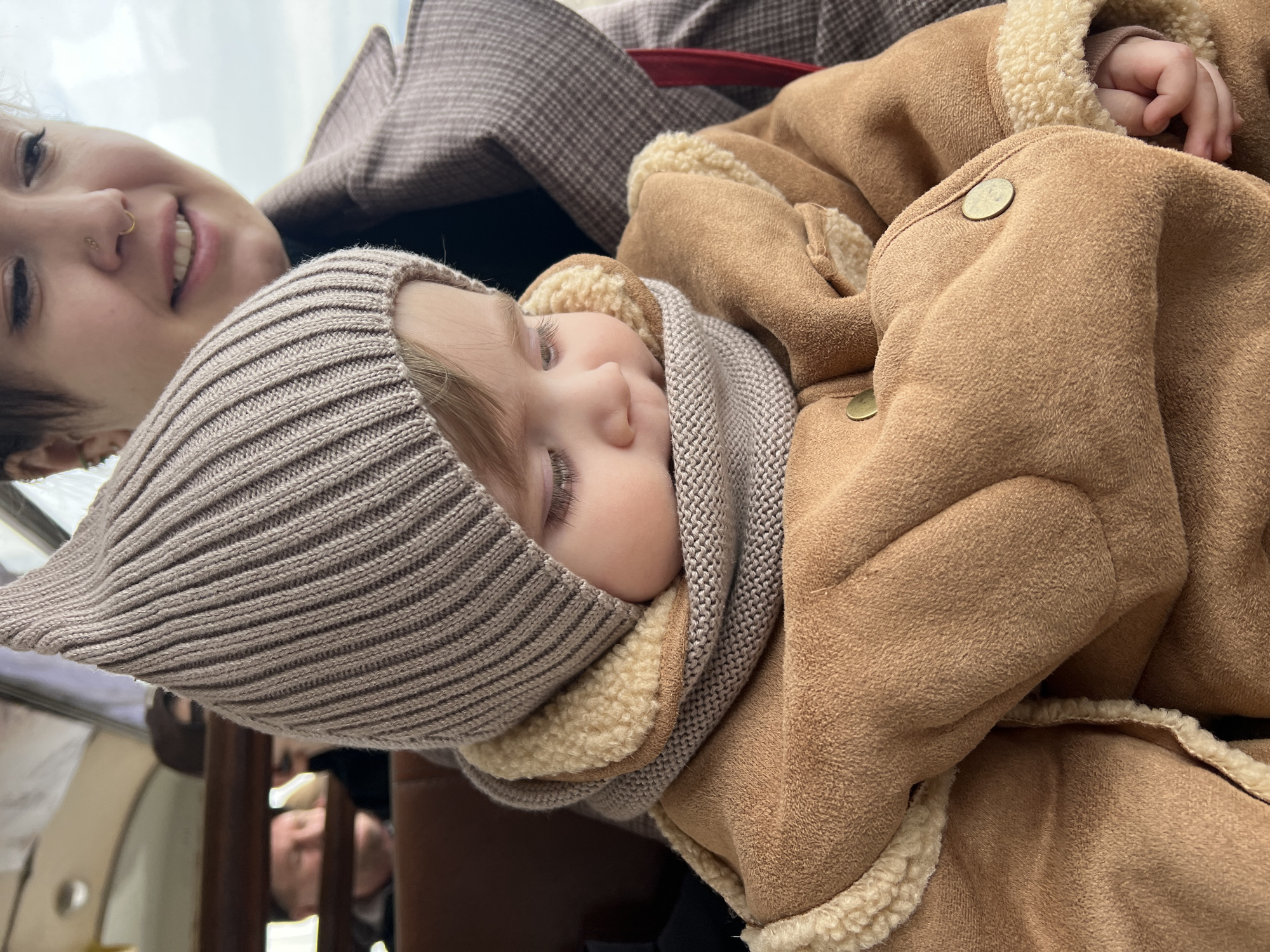 A photograph shows a baby wrapped in a brown jacket with a beige scarf and a light brown knit hat, with a woman visible near the baby's head. The location may be in the city of Chaikovsky, Russia. The baby is lying down, facing forward, with eyes closed. The woman is partially visible, smiling at the camera. The background is slightly blurred.