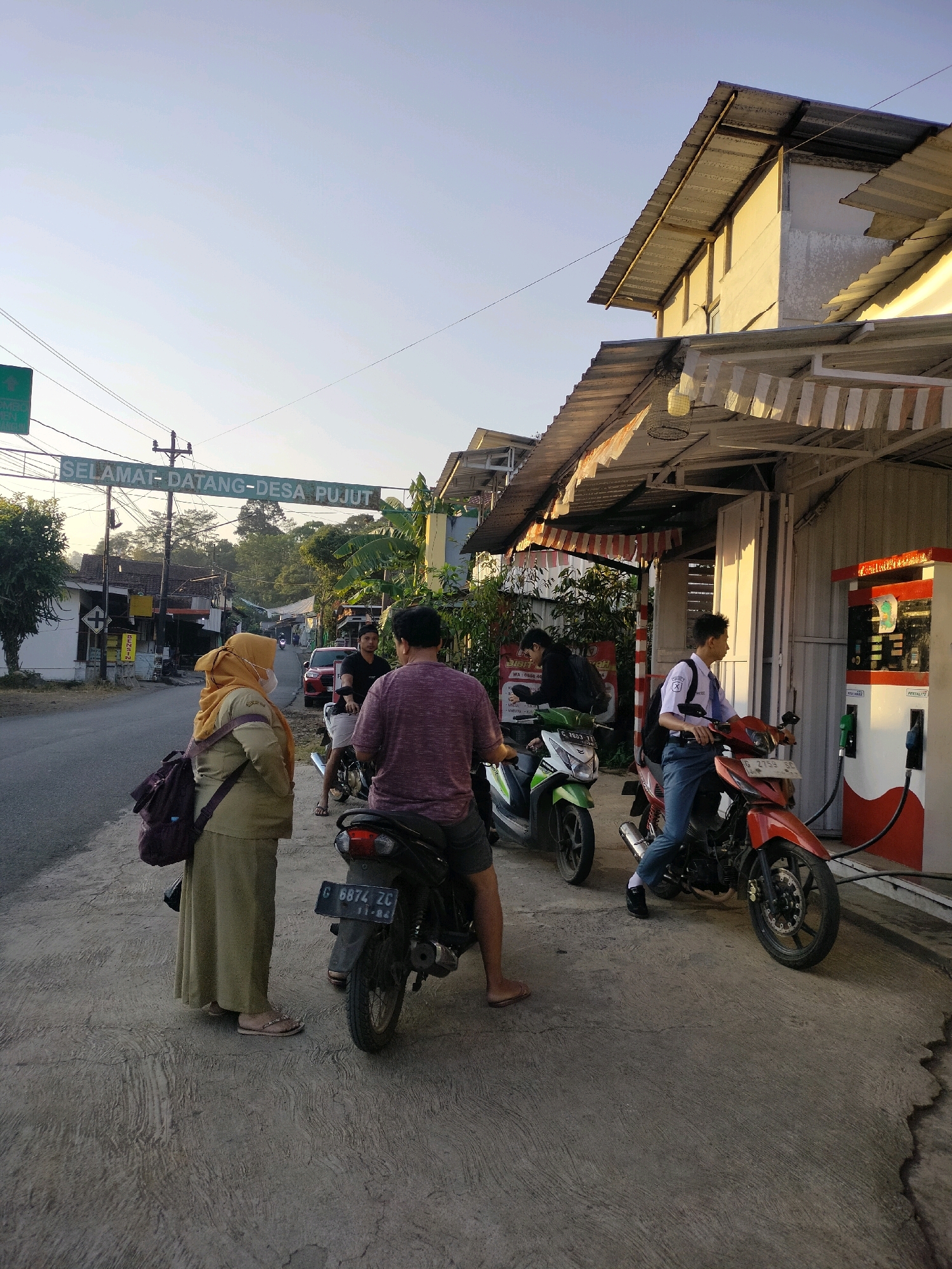 The image depicts a street scene in Tersono, Indonesia, near a gas station. A green sign with white lettering reads "SELAMAT DATANG DESA PUJUT," indicating it's the welcome sign for Pujut village. The foreground shows a concrete sidewalk with several individuals and motorcycles.
On the left, a woman wearing a light brown hijab and a long beige dress stands near a parked black motorcycle. She carries a dark purple backpack over her shoulder. To her right, a person in a purple t-shirt and grey shorts is seated on a black motorcycle. Another black motorcycle is visible in front of them.
Further to the right, a gas station with red and white pumps is situated alongside a building with a corrugated metal roof and an awning. Two young men on motorcycles are present at the gas station. One, wearing a white school uniform shirt and blue pants, sits on a red motorcycle, facing away from the camera. Another person, possibly a young man in a black t-shirt, is on a green motorcycle to his left, also facing away. A red car is parked further down the street on the left side.
The environment appears to be a rural or semi-urban area with trees and vegetation visible in the background. The lighting suggests it is late afternoon or early morning, with the sun casting a warm glow on the buildings. Power lines are strung across the street.