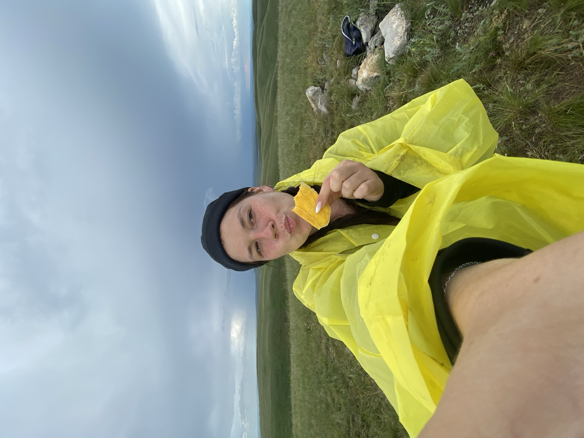 In Verkhneozernoe, Russia, a young woman wearing a yellow raincoat and a black beanie is captured in a selfie. She is holding a piece of yellow food, possibly cheese or fruit, to her lips and making a kissing face. The background depicts rolling green hills under a cloudy sky, suggesting an outdoor, possibly rural or natural setting. Rocks and a dark blue bag are visible on the ground to her right. The woman's left arm and part of her leg are visible in the foreground.