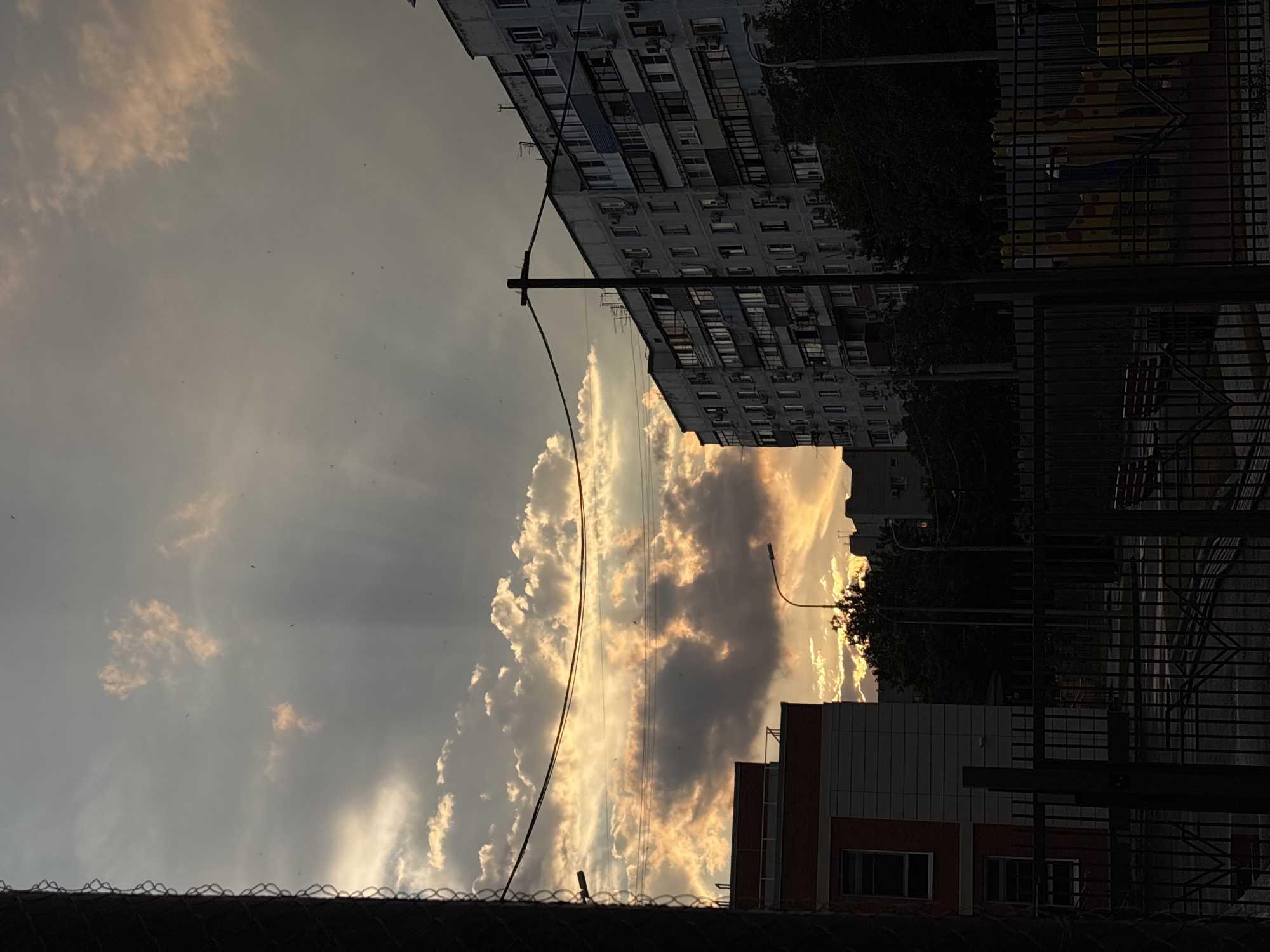 The image depicts an urban scene in Krasnodar, Russia, likely during sunset. The perspective is vertical, with the sky occupying a significant portion of the frame. Dramatic clouds, illuminated by the setting sun, create a play of light and shadow across the sky. Rays of sunlight pierce through gaps in the clouds, casting beams of light.
In the foreground, a dark, metallic fence runs horizontally across the bottom of the image, its intricate pattern adding texture. Behind the fence, a low-rise building with orange and white facades is visible, featuring windows. Further back, a tall, grey apartment building dominates the upper portion of the frame, its numerous windows and balconies suggesting a densely populated residential area. Silhouetted trees and a lamppost are also present, adding to the urban landscape. The overall mood is serene and perhaps a bit melancholic, characteristic of a fading day. The presence of multiple electrical wires crisscrossing the scene highlights the urban infrastructure. No text is visible in the image.