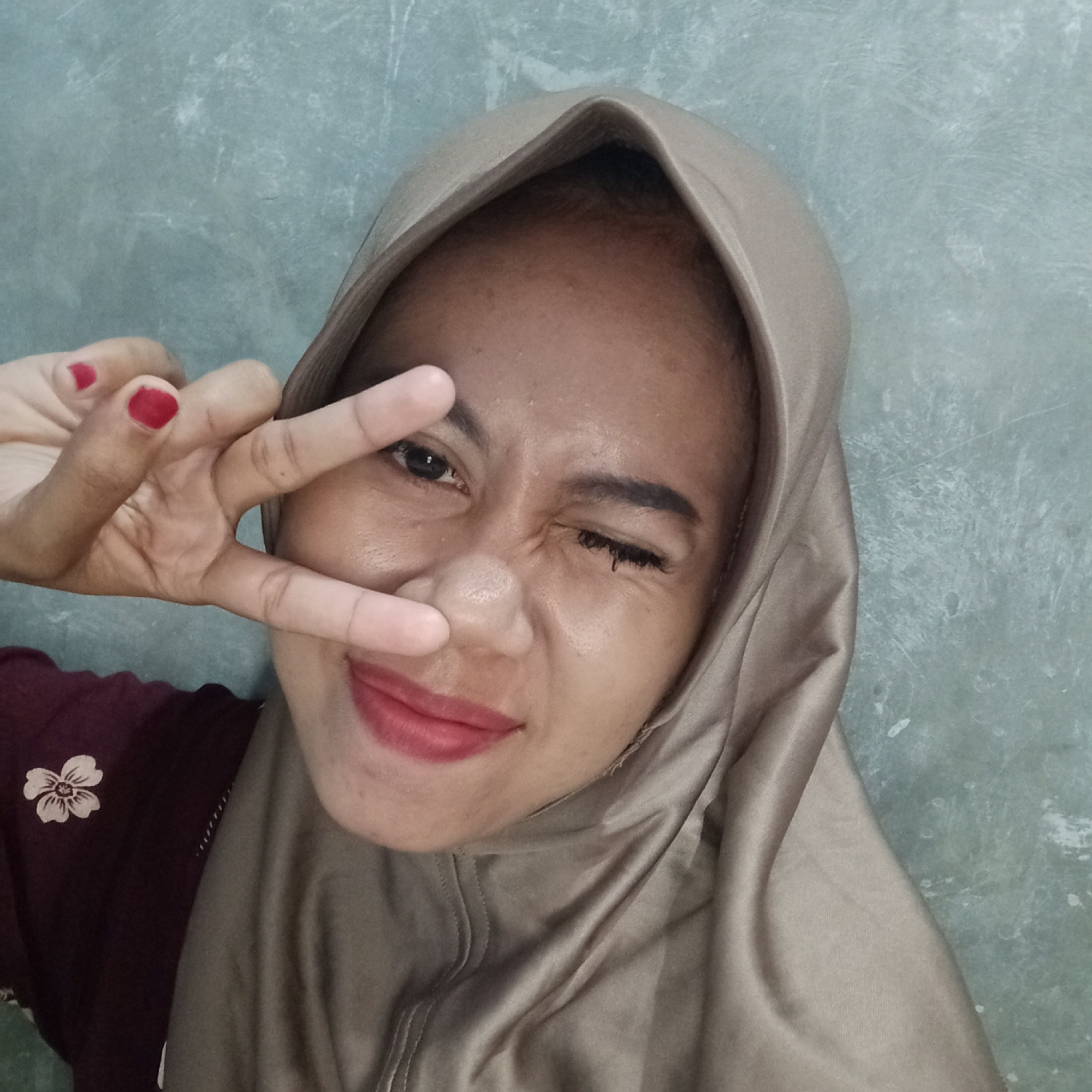 A young Indonesian woman, wearing a light brown hijab and a maroon shirt with a floral design, poses for a selfie against a grey, textured wall. She is positioned in the center of the frame, with her face slightly angled to the left. Her right eye is closed in a wink, while her left eye is open and looking towards the camera. She is holding her right hand up, with her index and middle fingers extended in a peace sign, and her thumb touching her ring and pinky fingers. Her lips are painted with a vibrant red lipstick. The lighting suggests an indoor setting, possibly a home or a casual public space. The image appears to be taken at close range, capturing the woman's facial expression and gesture. The background is plain and shows no distinguishing features that would indicate a specific location within Tegowanu Kulon, Indonesia.