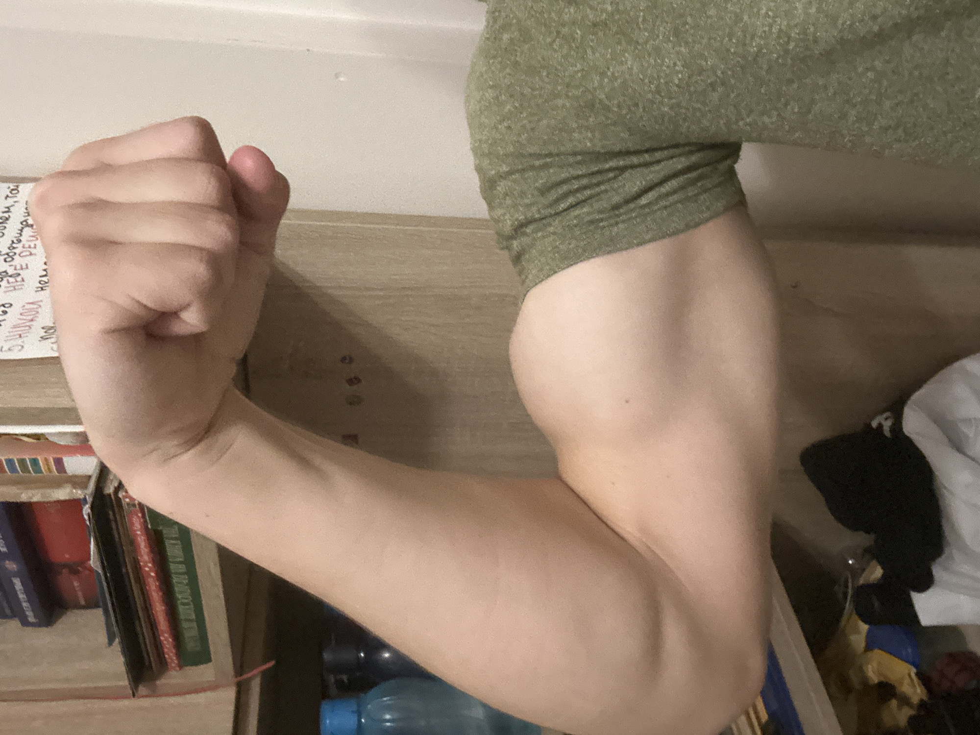 A close-up, indoor shot captures the right arm of an individual, flexed at the elbow with the hand clenched into a fist, prominently displaying the biceps muscle. The arm has light-toned skin with minor imperfections. The individual wears a speckled olive green short-sleeved t-shirt on the upper arm. The background reveals a domestic setting with light-colored wooden shelving units. On the upper shelf, multiple stacks of books are visible, including one with a red spine and another green-spined book partially showing Cyrillic text. A red cord or ribbon hangs vertically on the left. To the right of the arm, a light-colored wall features handwritten Cyrillic text, including the phrase "5. НИКОГДА НЕ РЕШ..." (5. NEVER DECIDE...). Below the arm, on a lower shelf or surface, two plastic bottles (one with a blue cap) and various out-of-focus items, including a black beanie and white fabric, are visible, suggesting a personal living space.