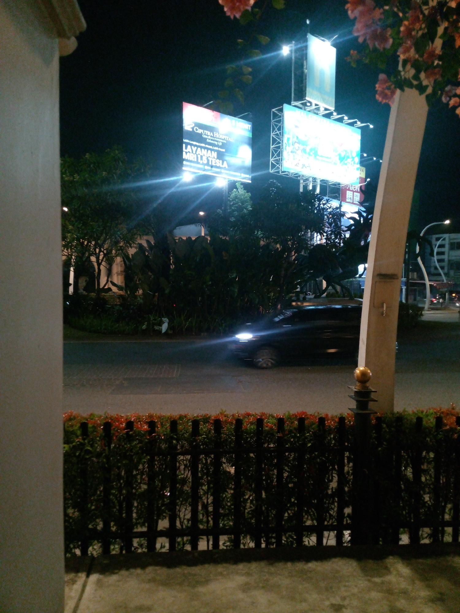 The image depicts a nighttime street scene in Bitung Jaya, Indonesia. A dark-colored car is driving on the wet asphalt road, its headlights illuminating the environment. A black metal fence with red-topped bushes is in the foreground. Behind the fence, trees and foliage are visible. Two large billboards are illuminated, one advertising "CIPUTRA HOSPITAL" and "LAYANAN MRI 1.5 TESLA," while the other displays a colorful graphic. A white pillar, part of a structure, is on the right side of the image. A street light and buildings are visible in the background. The overall lighting is dim, with artificial lights from the billboards and street lamps creating bright spots and reflections.