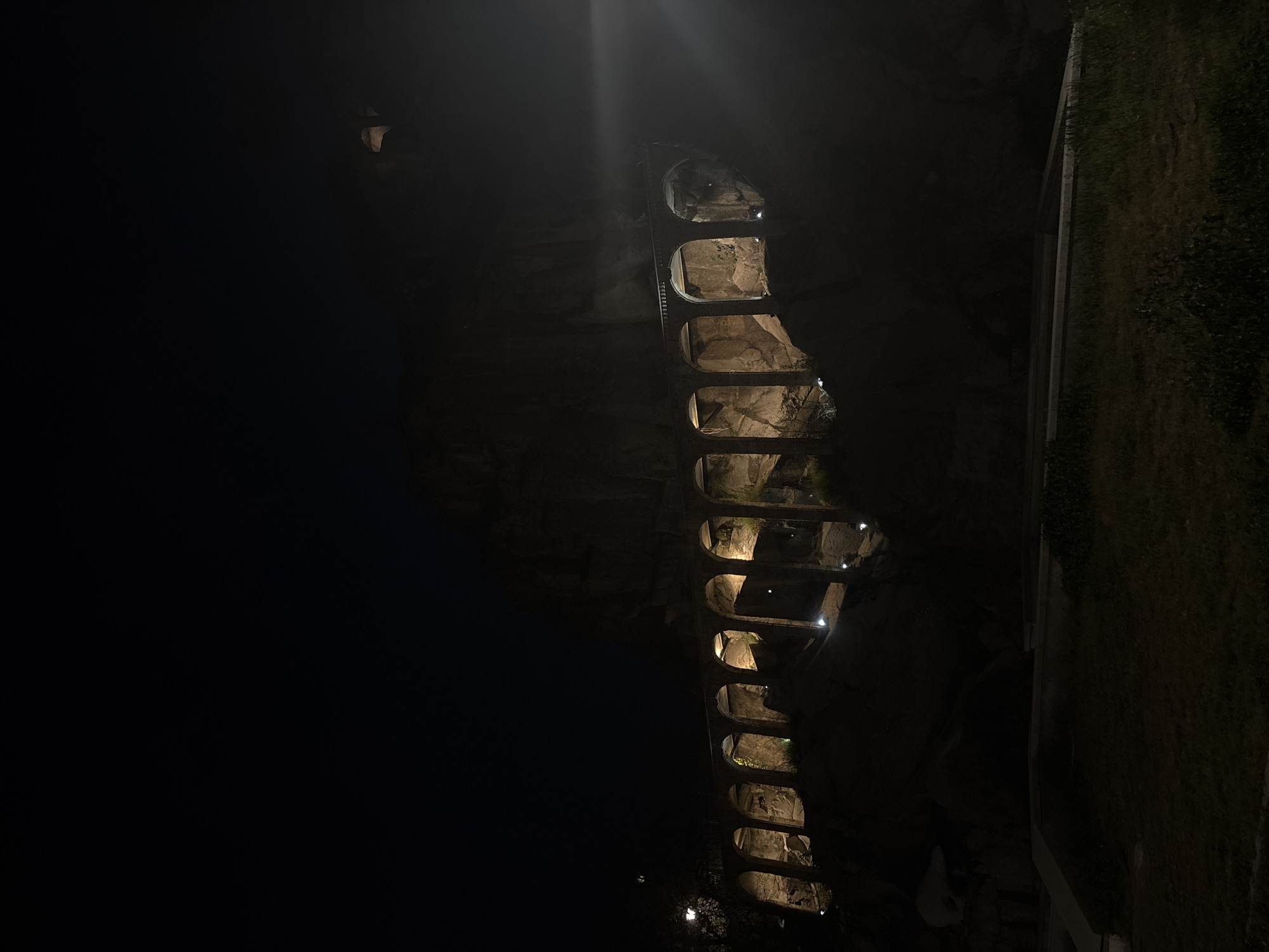 The image shows the Roman aqueduct in Plovdiv, Bulgaria at night. The illuminated arches of the aqueduct are visible against the dark sky and rocky background. To the right, there's a grassy area. There are no people visible.