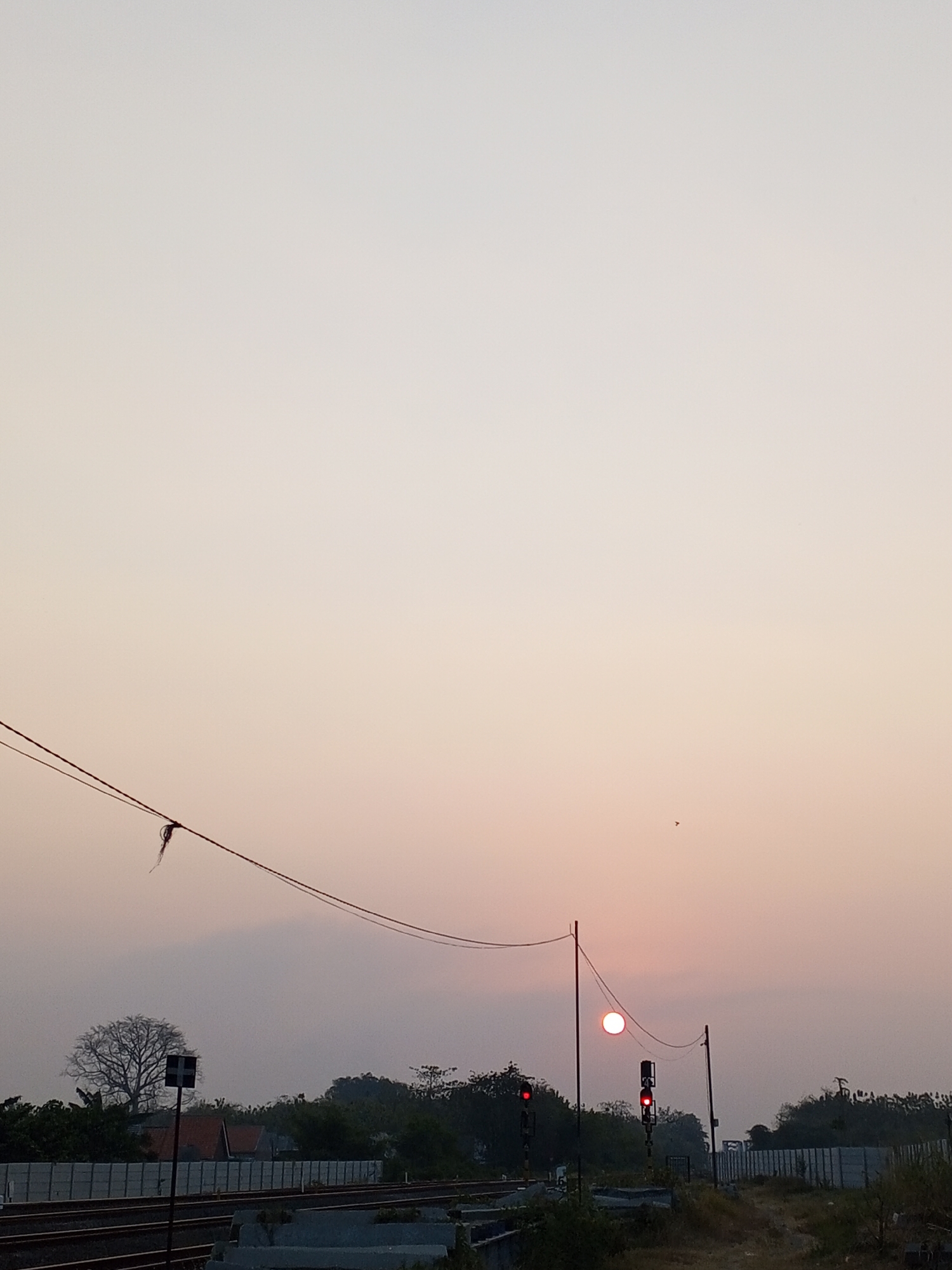 A serene, dusky scene unfolds in Pakusamben, Indonesia, as the sun dips towards the horizon, casting a warm, orange glow across the sky. The sky itself is a gradient of pale yellow and soft pink, creating a tranquil atmosphere.
In the foreground, train tracks stretch horizontally, hinting at the passage of trains through this rural Indonesian landscape. On the left, a silhouetted tree stands bare against the sky, its branches reaching upwards like intricate drawings. A black and white signpost with a cross symbol is positioned near the tree, possibly indicating a railway crossing.
Further to the right, two railway signals stand tall, their red lights glowing, suggesting a stop or caution for any approaching trains. The sun, a bright orange orb, is partially obscured by the signal on the right, its light diffused by the atmospheric haze. A solitary bird is visible as a small dark speck against the vast sky.
A line of trees forms a natural border in the middle ground, separating the railway from a distant row of concrete structures. The overall impression is one of quiet solitude, a peaceful moment captured at the edge of day in this Indonesian setting.