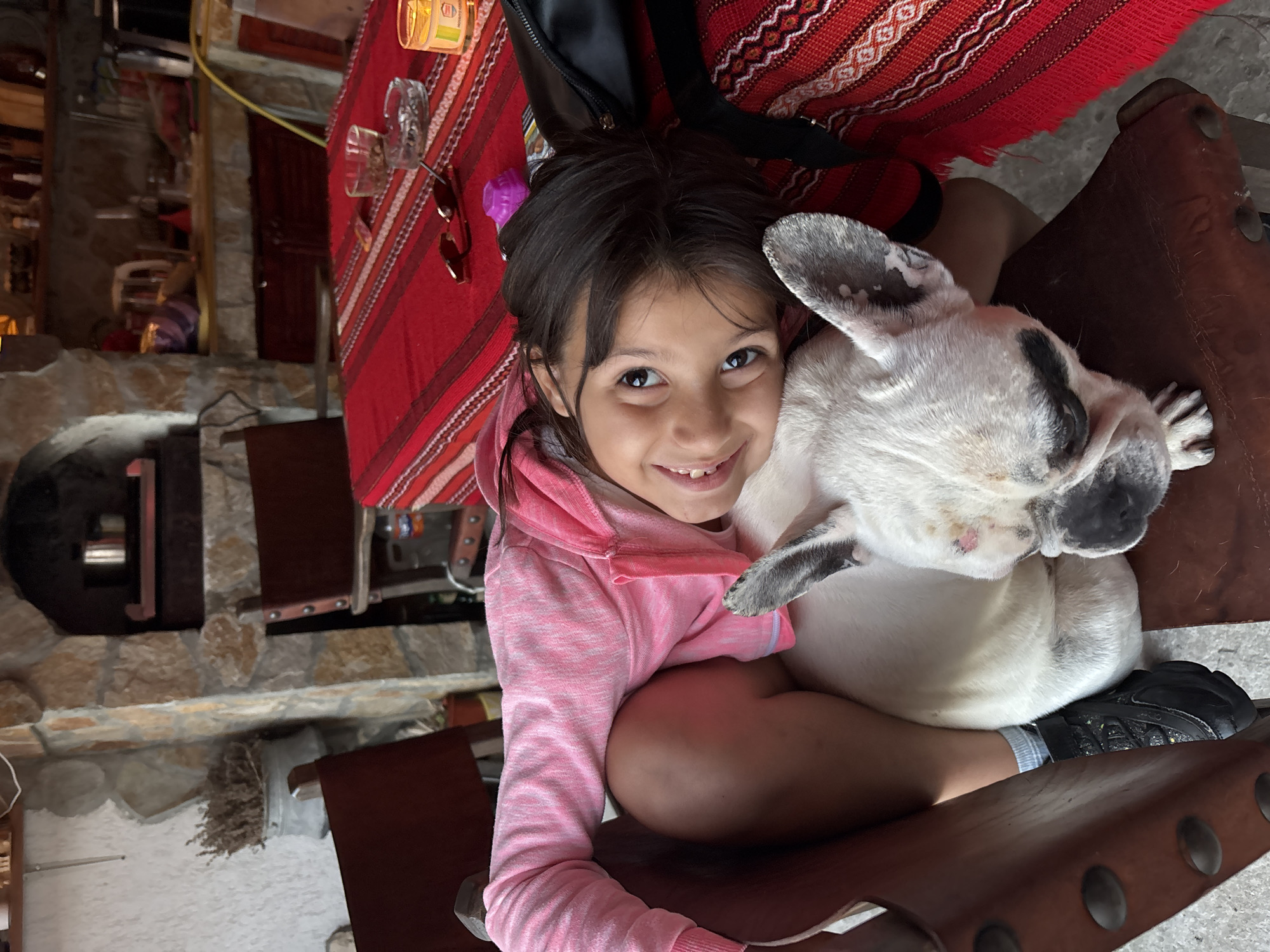 In the city of Enevo, Bulgaria, a young girl with dark hair, wearing a pink hooded sweatshirt, is lying down and resting her head on a white French bulldog. The girl is smiling and looking directly at the camera. The dog appears to be lying down as well, with its head resting on a brown leather chair. The scene appears to be outdoors, possibly on a patio or in a garden area, with stone walls and a fireplace in the background. A red and white striped tablecloth covers a table to the right, with sunglasses and a small candle on it.