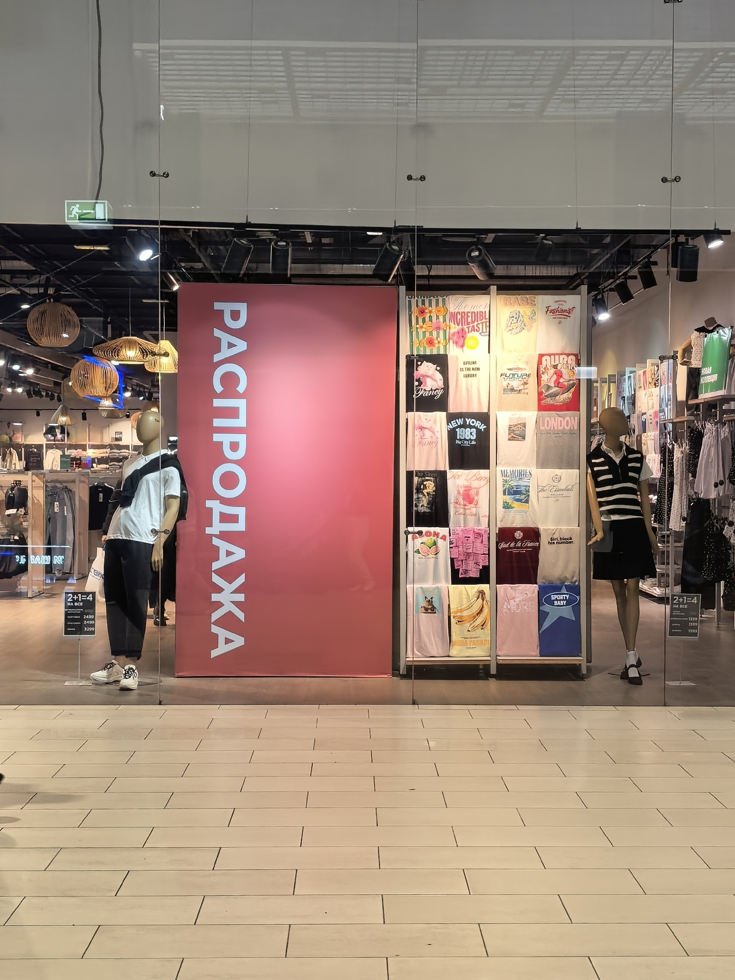 This image depicts the storefront of a clothing store, likely a fashion boutique or a chain retailer, with a prominent "SALE" sign in Russian, "РАСПРОДАЖА". The store appears to be located in a shopping mall or a commercial street in Ufa, Russia, judging by the Cyrillic signage and common retail aesthetics.
The entrance is framed by large glass panels, offering a clear view into the store's interior. On the left, a mannequin is displayed wearing a white t-shirt with a black diagonal stripe and dark pants, accessorized with white sneakers. To the right of the mannequin, a large red banner with white Cyrillic lettering announces the sale.
In the center of the display, a rack showcases an assortment of graphic t-shirts, each featuring different designs and slogans, such as "INCREDIBLE TASTE," "BABE," "Fashionist," "NEW YORK 1983," "LONDON," and others. These t-shirts are neatly folded and arranged in rows.
To the right of the t-shirt display, another mannequin stands, dressed in a black and white striped sleeveless top and a black skirt, paired with white socks and black shoes. Behind this mannequin, racks of clothing are visible, including dresses and blouses, suggesting a varied inventory.
The store's interior is well-lit with modern track lighting. Decorative pendant lights made of woven material hang from the ceiling, adding a touch of warmth and style to the ambiance. The floor is tiled with light-colored rectangular tiles, reflecting the overhead lighting. An "EXIT" sign with a green running figure is visible on the glass panel above the left side of the entrance.
In the foreground, a price promotion sign indicating "2+1=4" is visible on the glass, suggesting a buy-two-get-one-free or similar promotional offer. The prices listed are in Russian Rubles. The overall impression is of a modern, well-organized retail space focused on trendy apparel.