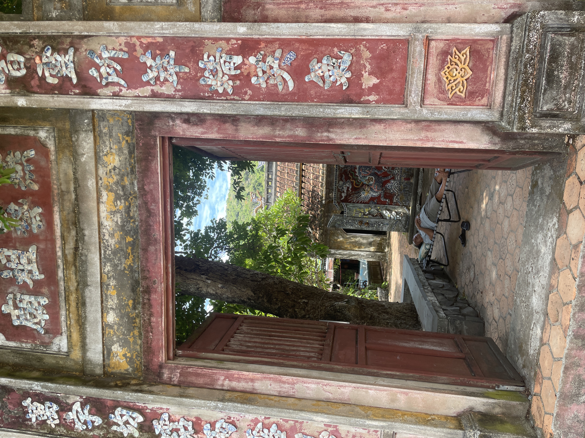 The image captures a view through an ornate red gateway, likely at the entrance to a historical site or temple in Đà Nẵng, Vietnam. The gateway is adorned with colorful mosaic characters. To the right of the entrance, a person is lying on a cot, resting. Beyond the gateway, a courtyard is visible with traditional Vietnamese architecture, including tiled roofs. A large tree stands prominently within the courtyard. The overall scene suggests a tranquil and historical setting.