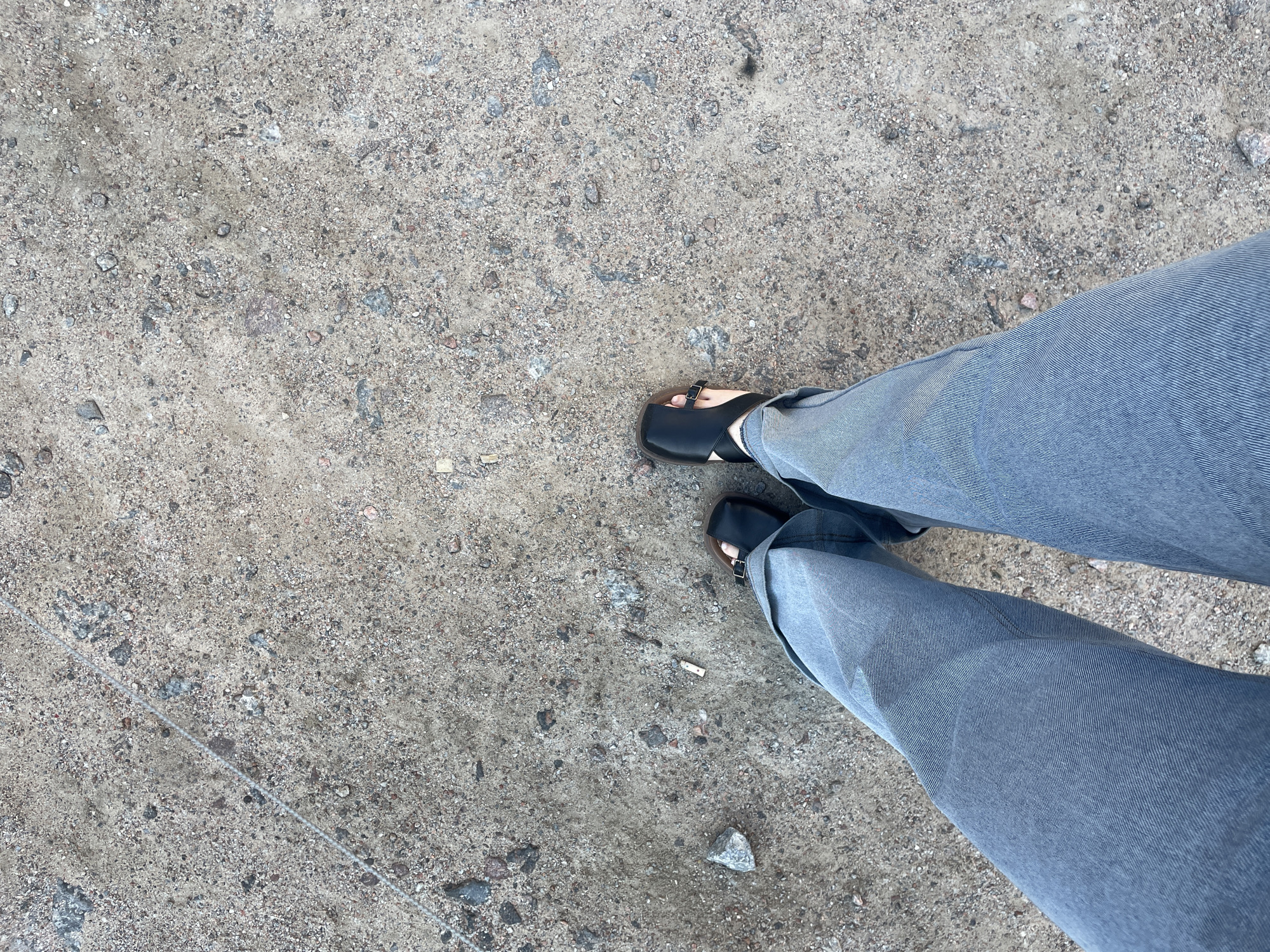 The image shows a top-down view of a person's legs wearing loose-fitting grey pants and black sandals, standing on a ground composed of sand and small rocks. A thin, light-colored string is visible diagonally across the upper left portion of the frame. There is a small, white, cylindrical object resembling a cigarette butt on the ground near the person's right foot. The environment appears to be outdoors, possibly a pathway or an unpaved area within a city. The lighting suggests it is daytime.