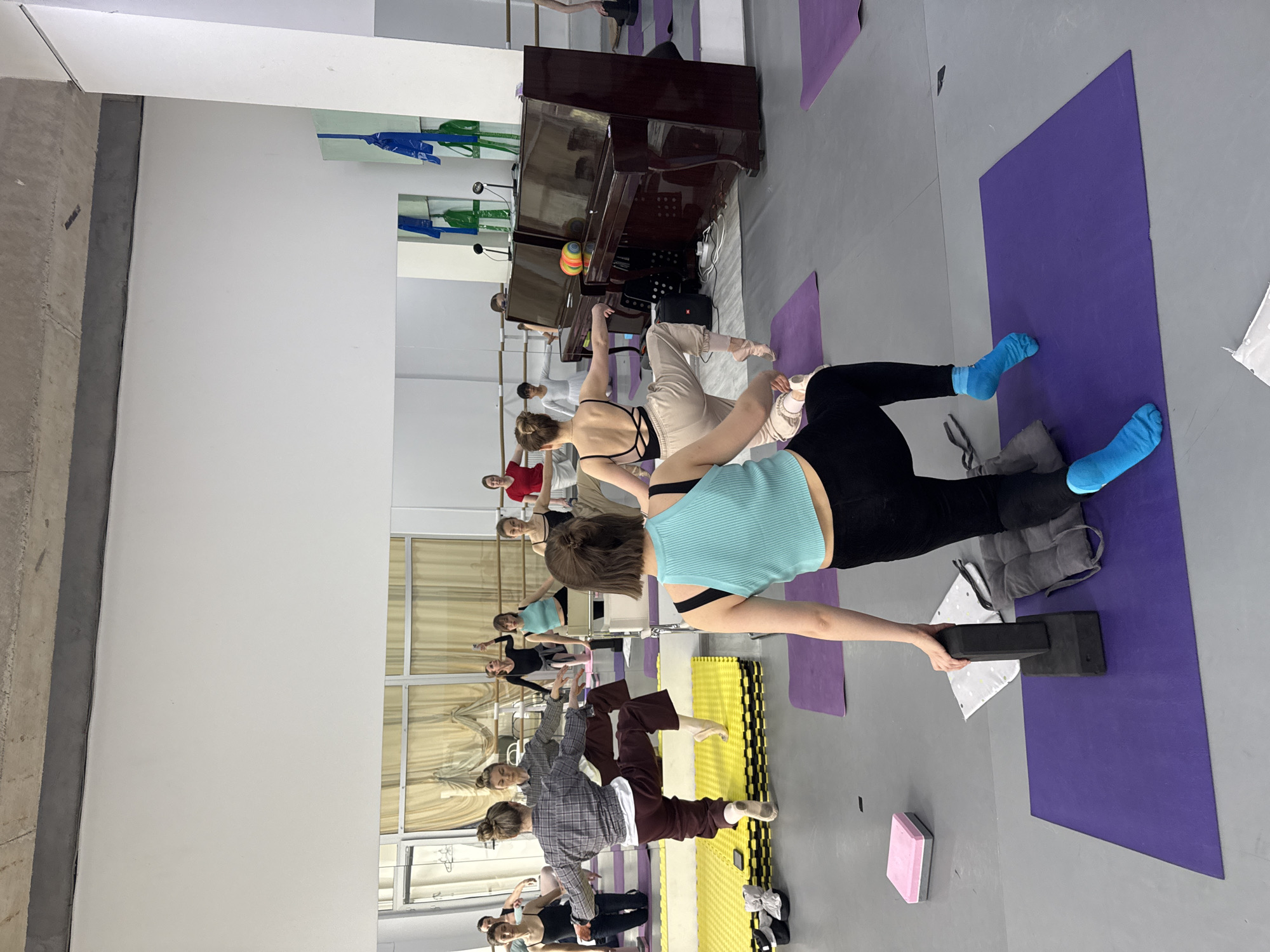 The image captures an indoor scene within what appears to be a dance or fitness studio. A group of women, primarily young adults, are present, engaged in various poses and stretches, suggesting a class or practice session.
In the foreground, a woman wearing a light blue ribbed top, black leggings, and bright blue socks is prominent. She is utilizing a black yoga block on a purple yoga mat, possibly for balance or a stretching exercise, with her arm extended to support herself on the block. Next to her on the mat is a grey cushion and a small white patterned cloth. Another pink and grey yoga block is visible on the floor further left.
Behind her, other individuals are visible. One woman in a black top and light-colored pants is near a dark wooden upright piano, which has sheet music and two microphones on it. Reflections in the large wall-mounted mirrors show more people, some appearing to use a ballet barre. The studio has a light grey floor, white walls, and a concrete support beam on the left. Windows with light-colored curtains are reflected in the mirrors, indicating external light sources.
The overall atmosphere suggests a focused and active environment, likely a dance or yoga class. The time of day is not explicitly clear but the studio is well-lit. No discernible text is visible in the image.