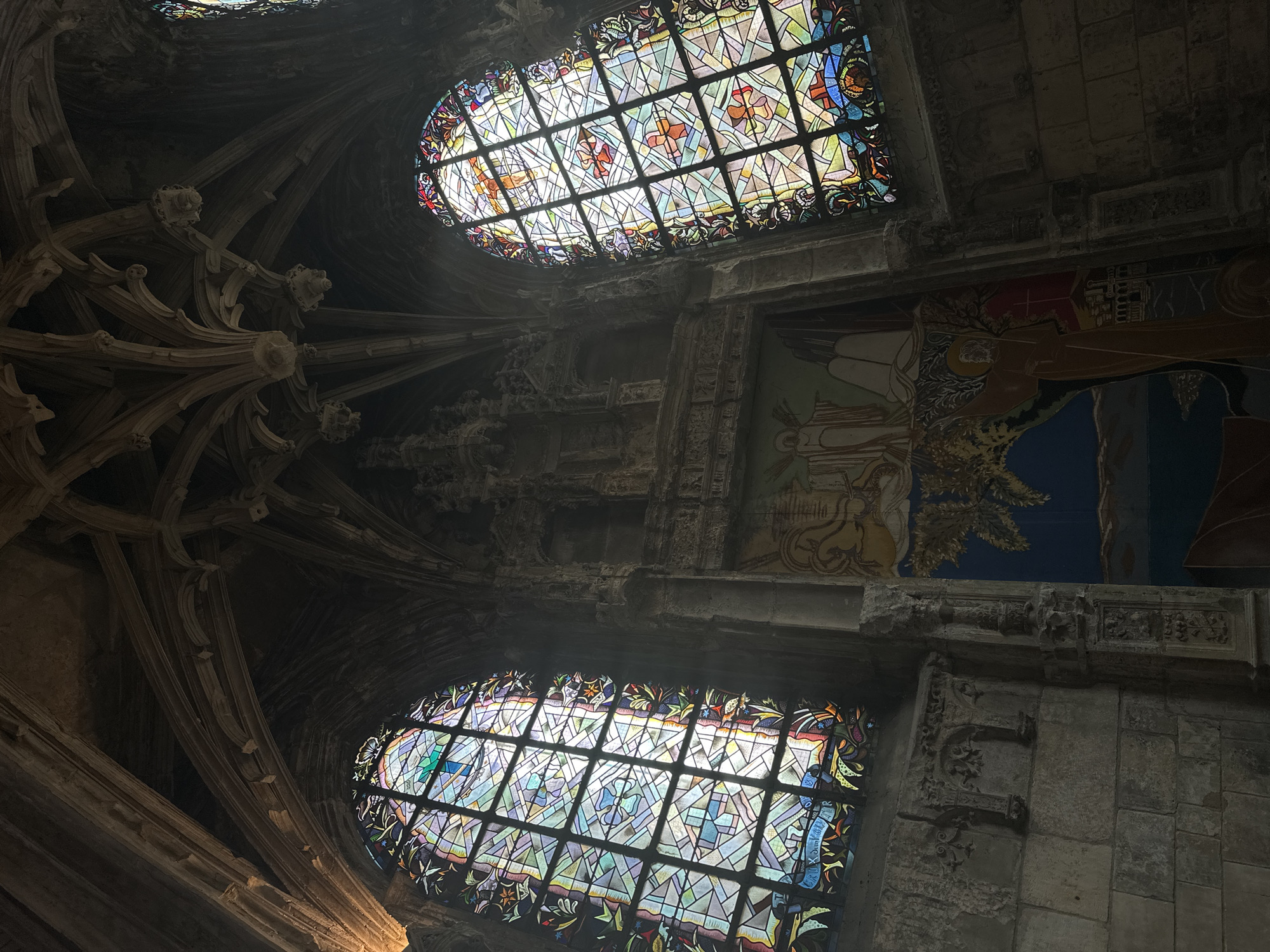 The image shows the interior of the Church of Saint-Jacques in Dieppe, France, featuring ornate Gothic architecture and stained glass windows. The ceiling exhibits intricate rib vaulting, with carved stone embellishments. Two large, arched stained glass windows are prominent, one on the left and one on the right, displaying colorful geometric patterns and figurative scenes. Below the right window, a large mural depicting religious imagery is visible, featuring figures and a landscape. The walls are constructed of rough-hewn stone blocks, with decorative stone carvings along the architectural elements. Sunlight streams through the stained glass, illuminating the dimly lit interior. The scene is devoid of people.