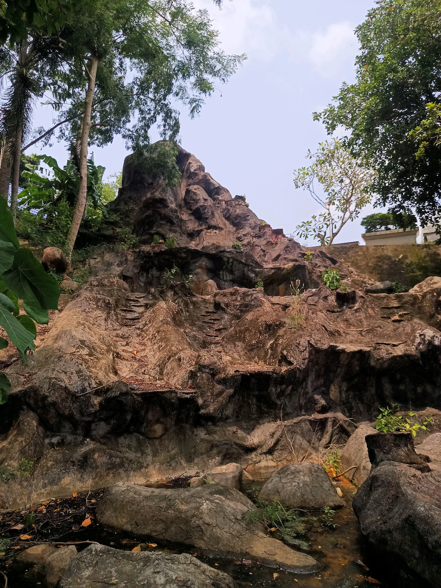 A man-made rock formation, possibly a waterfall or a garden feature, is the central focus of this outdoor scene in Sai Thai, Thailand. The rock formation is textured and appears to be made of concrete or similar material, designed to mimic natural rock. It has several levels and ledges, with some suggesting the presence of steps or pathways. Vegetation, including trees and smaller plants, grows around and on the rock structure, with some greenery visible at the base and on higher parts of the formation. There is a body of water at the bottom of the rock formation, appearing murky and reflecting the surrounding environment. In the upper left of the image, a portion of what appears to be a person, possibly a man, is visible behind the foliage and rocks. The sky above is overcast, indicating a cloudy day. The surrounding environment suggests a garden or park setting.
