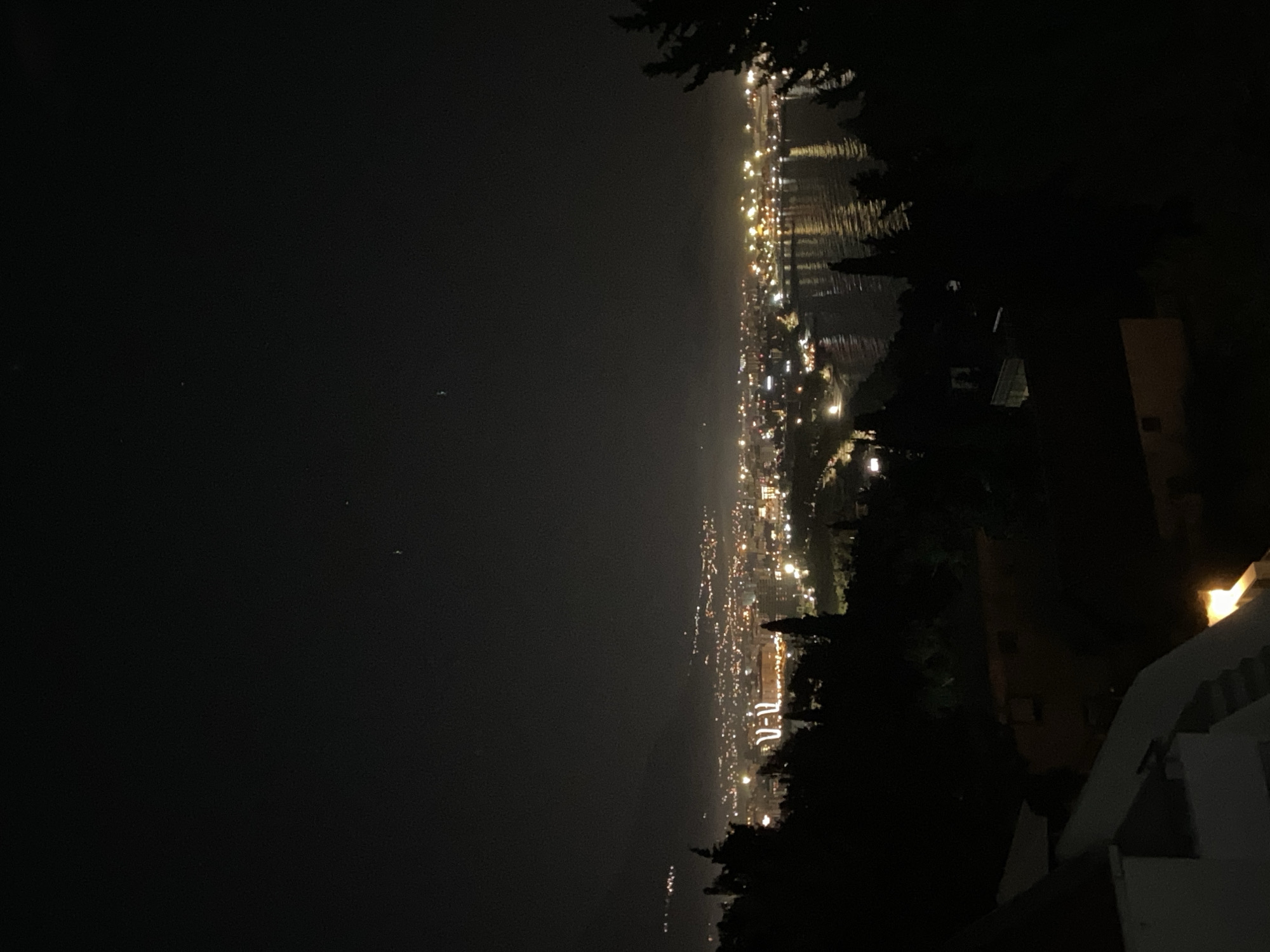 An outdoor, nighttime view, likely from an elevated position in Šušanj, Montenegro, reveals the city's lights stretching along the coastline. The lights create a bright strip against the dark sea and sky. Some buildings and trees are silhouetted in the foreground, providing a sense of depth. A partially illuminated building structure is visible in the lower right corner of the frame.