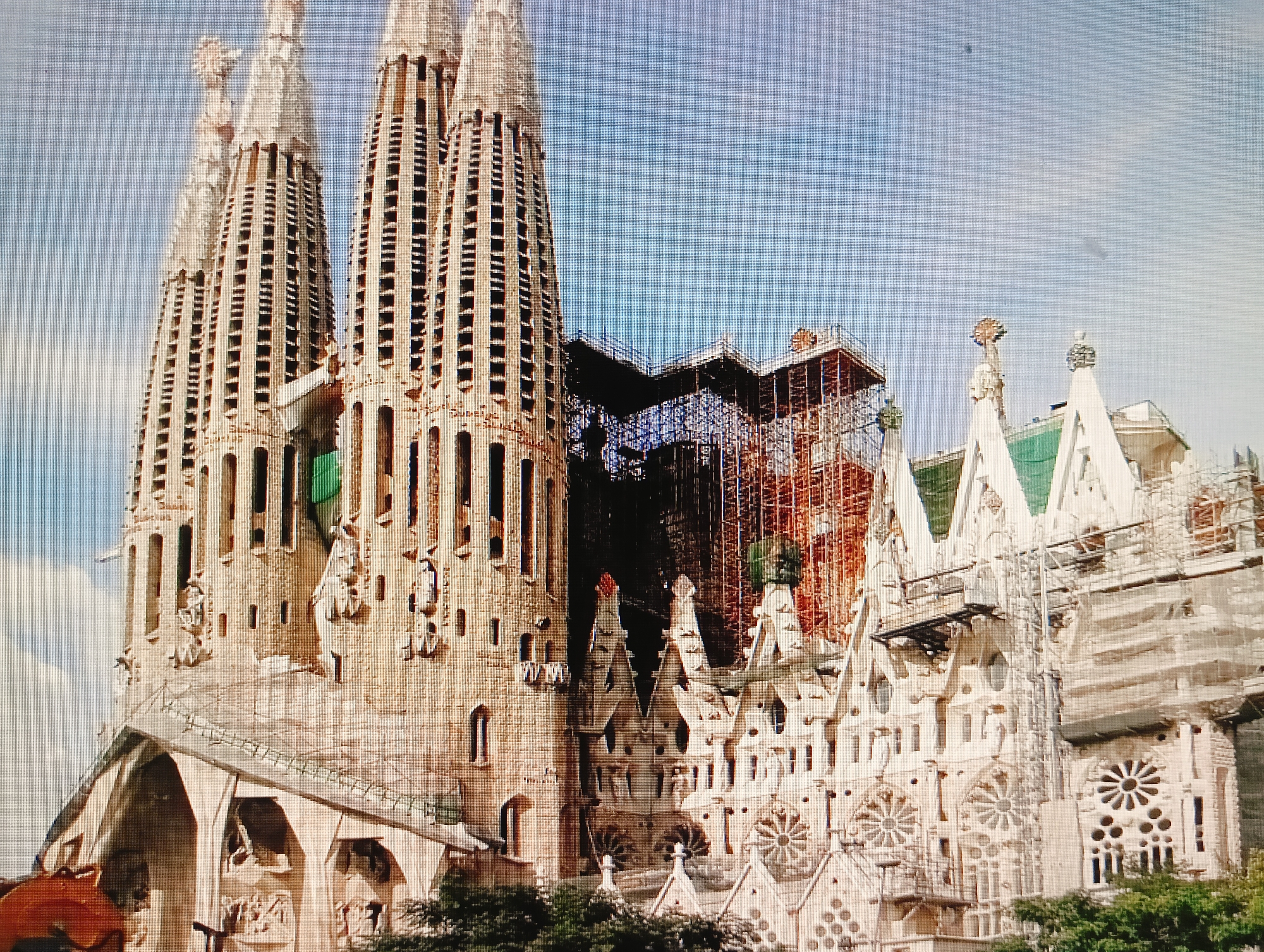 The image depicts a daytime exterior view of a large, ornate, stone building, likely a cathedral or basilica, under construction or renovation. The architecture is elaborate, with many spires, intricate carvings, and arched windows. Scaffolding is visible around some sections of the building, indicating ongoing work. The sky is a light blue with some clouds. Trees and greenery are visible in the foreground, suggesting an outdoor setting in a city. There are no people or discernable actions taking place. The building's construction and style are strongly reminiscent of Antoni Gaudí's work, specifically the Sagrada Familia in Barcelona, Spain. There is no indication in the image that this location is in Sluck, Belarus.