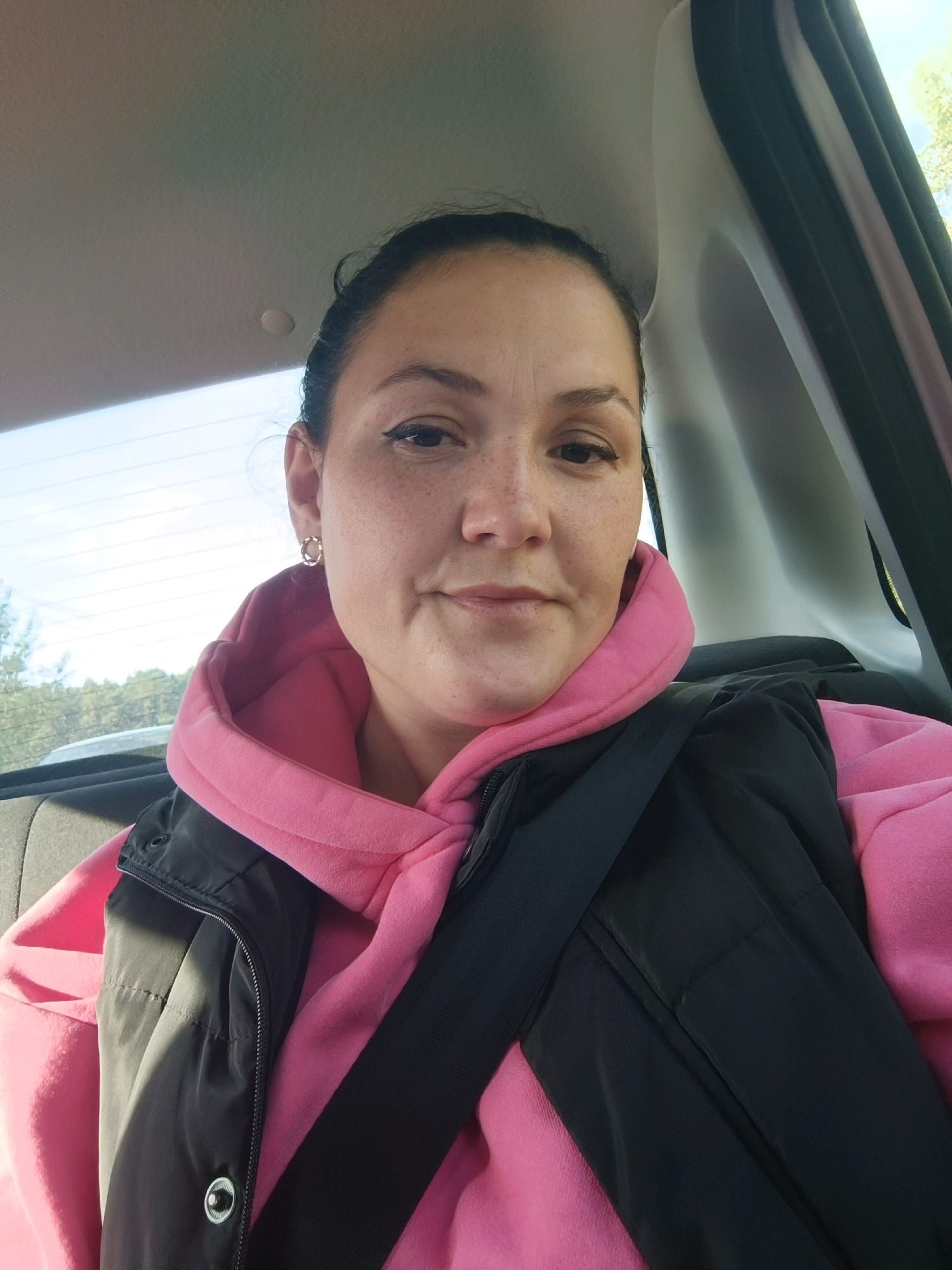 A close-up, waist-up shot shows a woman with dark hair pulled back and fair skin with freckles. She is wearing a bright pink hooded sweatshirt and a black vest over it, with a black seatbelt crossing her chest. Her left ear has a small, circular gold earring. The background suggests the interior of a car, with a window showing trees and a bright sky. No specific location in Chaikovsky, Russia, can be determined from the image.