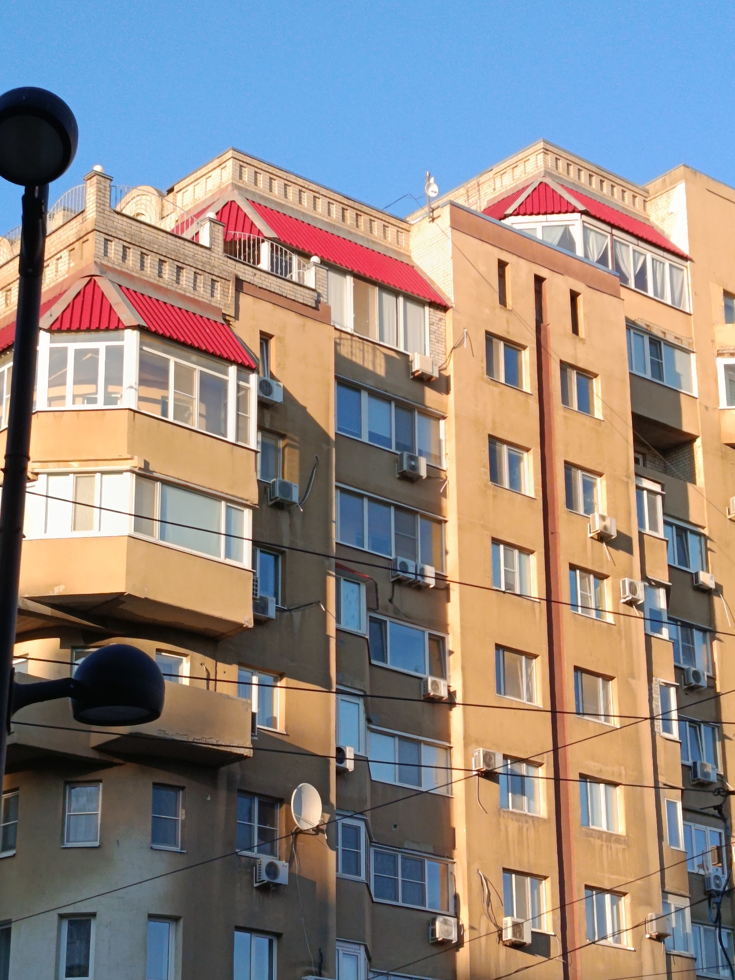 The image shows a multi-story apartment building in Saratov, Russia, under a clear blue sky. The building has a light brown facade with numerous windows and balconies. Many of the windows have air conditioning units mounted beneath them. The building features distinctive red-tiled gabled roofs on several sections, adding a touch of color to the architecture. A street lamp is visible on the left side of the frame, and electrical wires crisscross the building's facade. The overall scene depicts a typical urban residential environment.