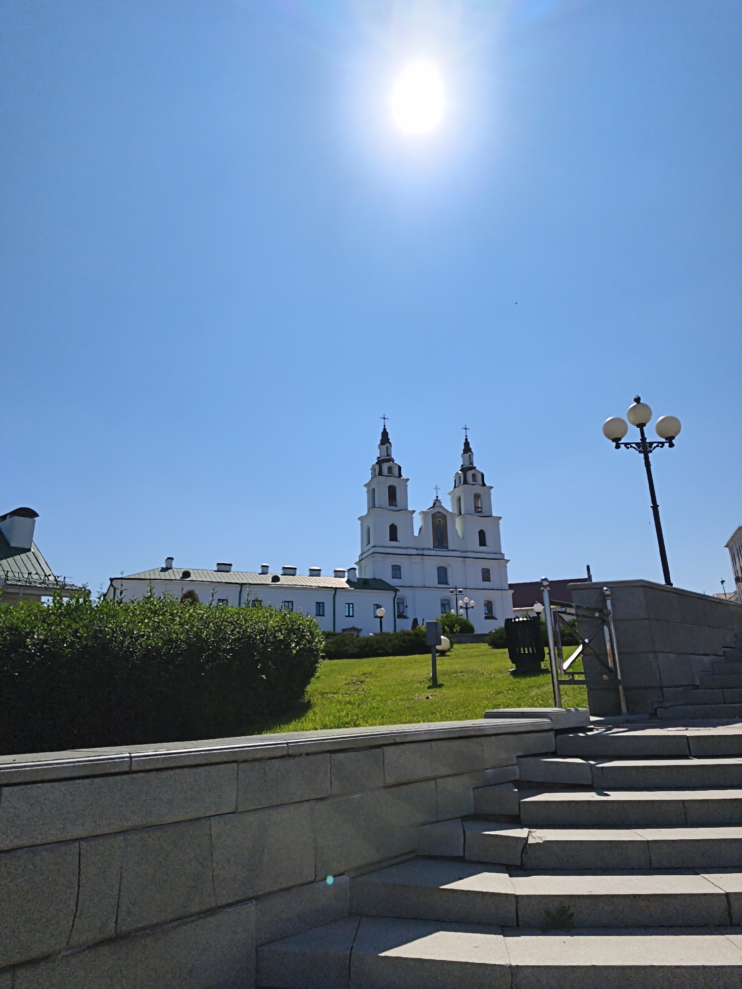 This is a ground-level, exterior, daytime, medium shot of the Holy Spirit Cathedral in Minsk, Belarus, under a bright, clear blue sky. The sun is shining brightly in the upper center of the frame, creating lens flare. The white, neoclassical cathedral with two prominent towers is situated on a grassy hill in the background. In the foreground, there is a stone wall and a set of concrete stairs. A black lamppost with three globes stands to the right of the cathedral. Lush green bushes are visible in front of the cathedral. The scene is quiet and appears to be a warm day.