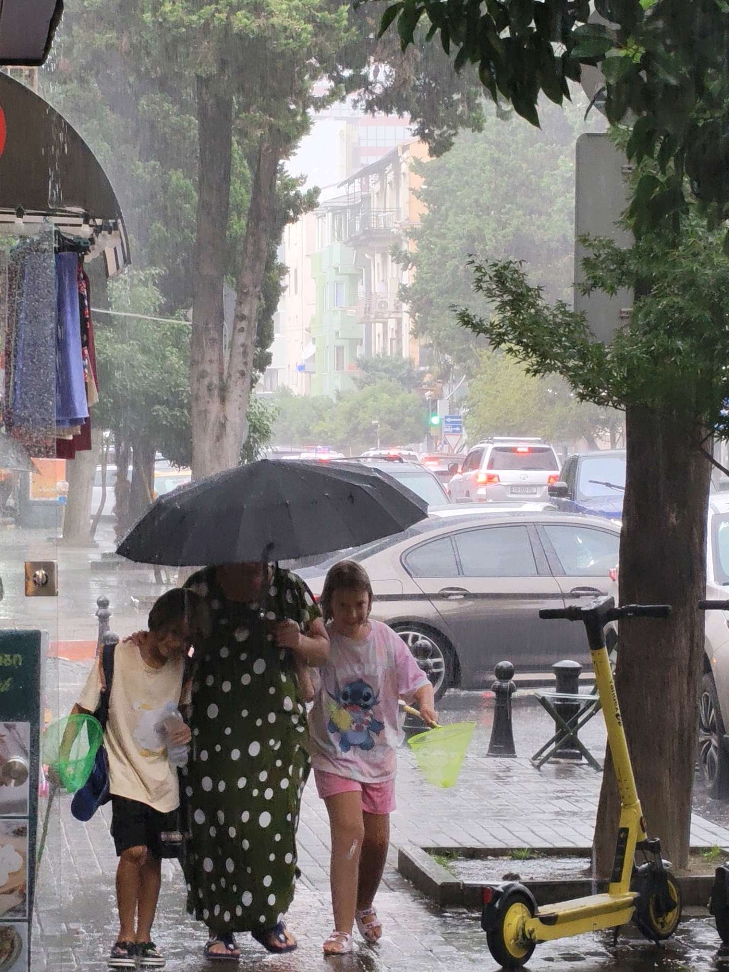 In Batumi, Georgia, during a heavy downpour, three individuals, a woman and two children, are walking under a large black umbrella. The woman, dressed in a dark green polka-dotted dress, stands in the center. To her left, a child wearing a cream-colored t-shirt and black shorts, with a green bag and a plastic bottle, walks with their arm around the woman. To the woman's right, another child in a pink t-shirt with a cartoon character and pink shorts carries a yellow net bag. They are on a wet, paved sidewalk next to a street with several cars, including a white SUV and a dark sedan. A bright yellow electric scooter stands to the right of the group, and trees and buildings are visible in the background. The weather is clearly rainy, with visible water droplets in the air and on surfaces.