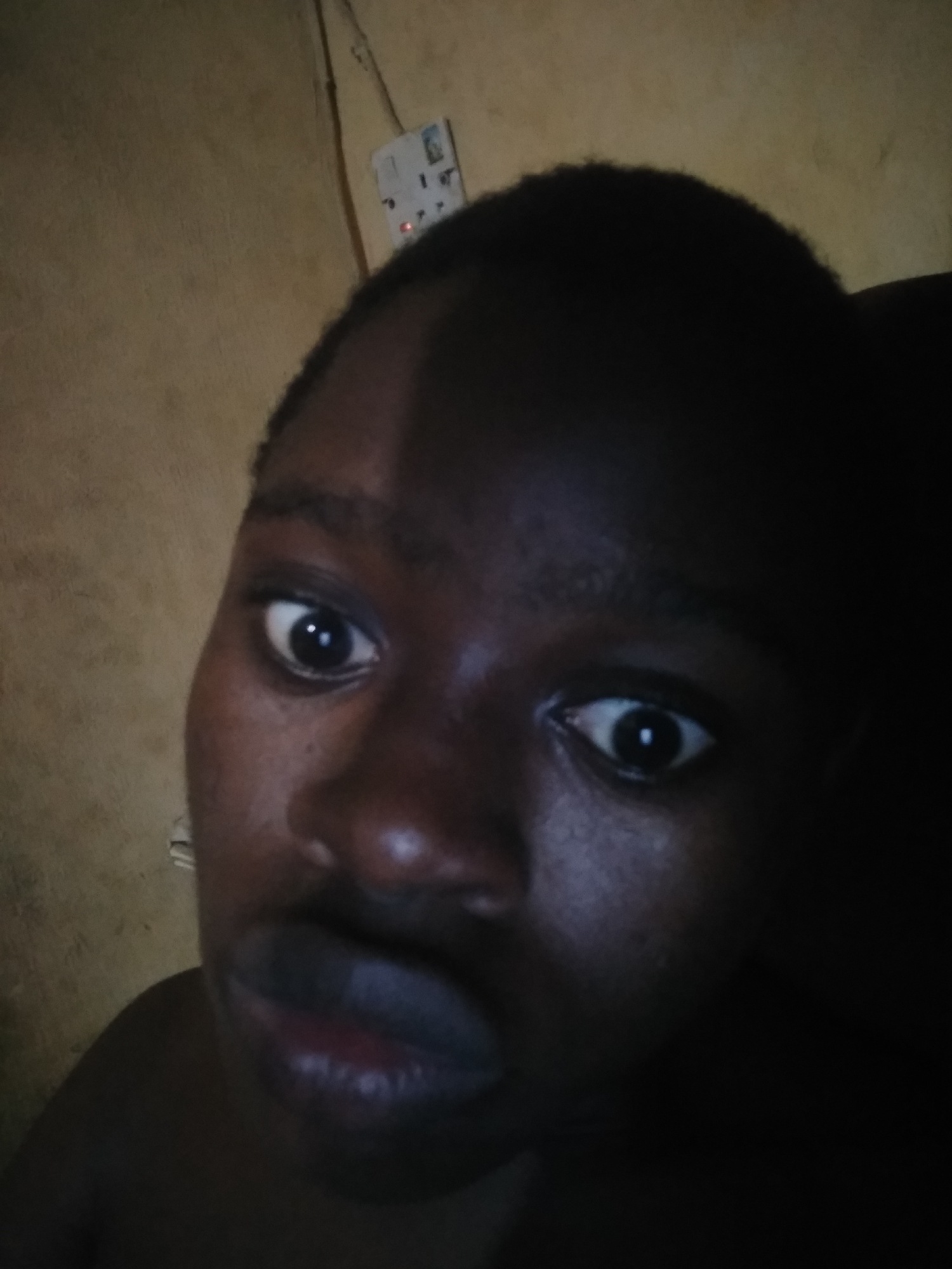 A close-up, low-angle indoor shot features a person with dark skin. The person's eyes are wide and directed slightly off-center. Part of the person's head is obscured by shadow, and the background consists of a wall and an electrical outlet. The city and precise location within it are not discernible from the provided image.