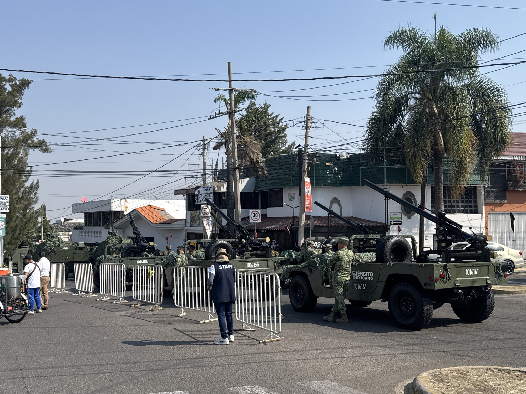 The image shows a street in the city of Puebla, Mexico. A line of military vehicles, identified by the text "EJERCITO MEXICANO" and license plates reading "1016161" on their rear, are positioned along the street, potentially for a parade or security detail. The vehicles are olive green and appear to have anti-aircraft guns mounted on them. Soldiers in camouflage uniforms are standing near the vehicles. A person wearing a blue vest with "STAFF" printed on the back stands behind a line of white metal crowd control barriers, seemingly overseeing the scene. Civilians are also present, with two men standing near a food cart on the left side of the image. The background includes buildings, trees (including palm trees), utility poles with overhead wires, and street signs. One sign shows a speed limit of 30 km/h.