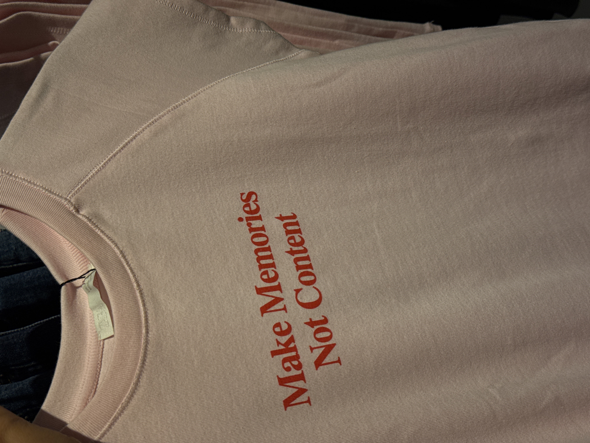 The image shows a close-up of a pink sweatshirt with the text "Make Memories Not Content" printed in red. A tag is visible near the neckline. The environment consists of other folded pink sweatshirts and a dark denim-like material beneath the pink sweatshirt in focus. The city location, Kuta, Indonesia, is not evident in the provided image, as no recognizable landmarks or contextual details point to this specific location. Therefore, the description is limited to the visible elements within the image.