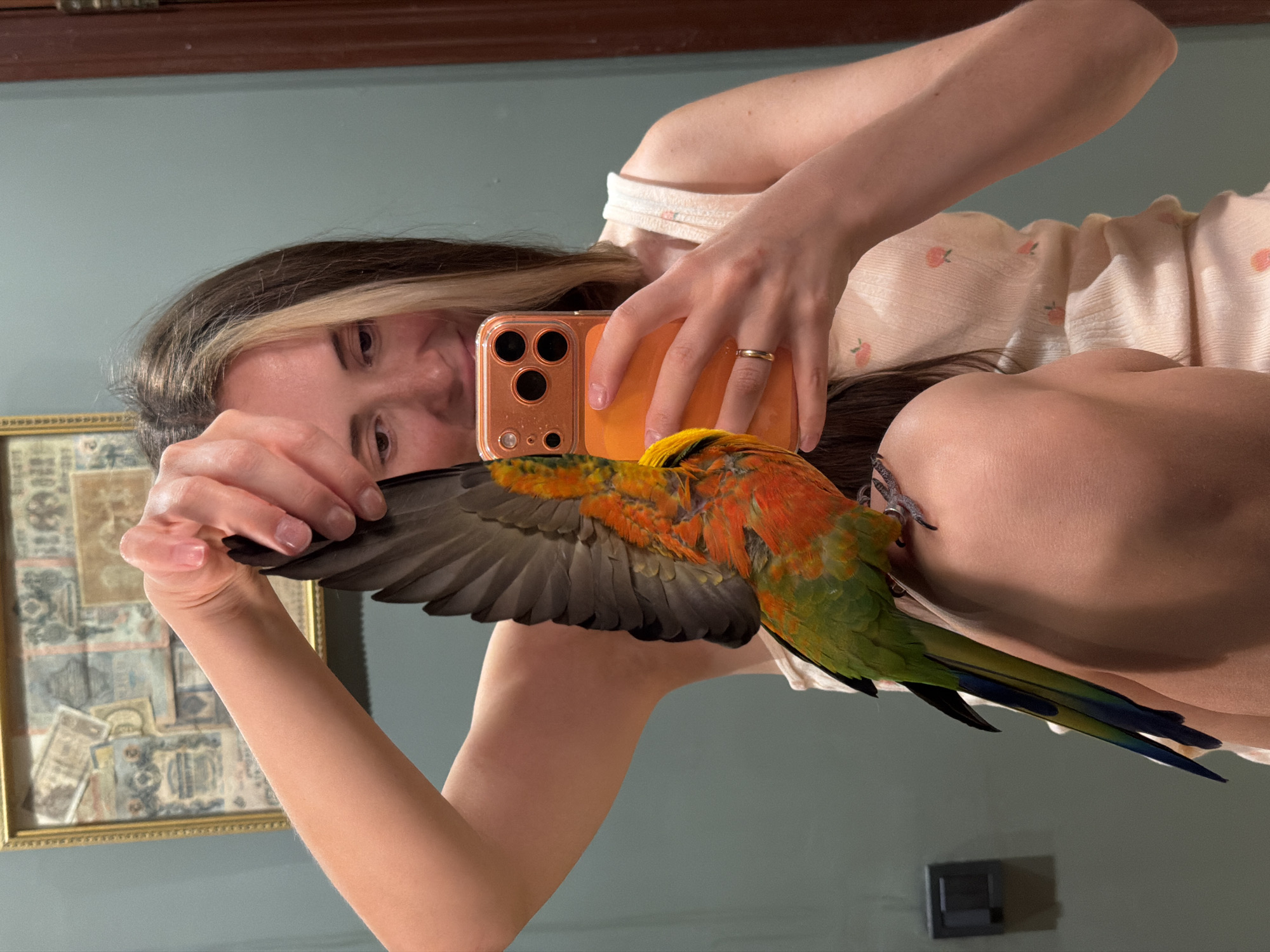 A young woman is taking a selfie in a bathroom. She has long brown hair with blonde streaks and is wearing a light-colored tank top with a fruit pattern. A brightly colored parrot, with green, orange, and yellow plumage, is perched on her right arm, its wing extended. The woman is holding an orange smartphone in her right hand, angled to capture the reflection. The background shows a pale green wall with a framed artwork, possibly old currency or stamps, on the left and a wooden door frame on the right. The woman appears to be looking at the phone screen, with a slight smile. The parrot's talons are visible gripping her arm. The overall lighting suggests an indoor environment, possibly a home bathroom. The provided metadata does not contain any information about the location being in the city of Khimki, Russia.