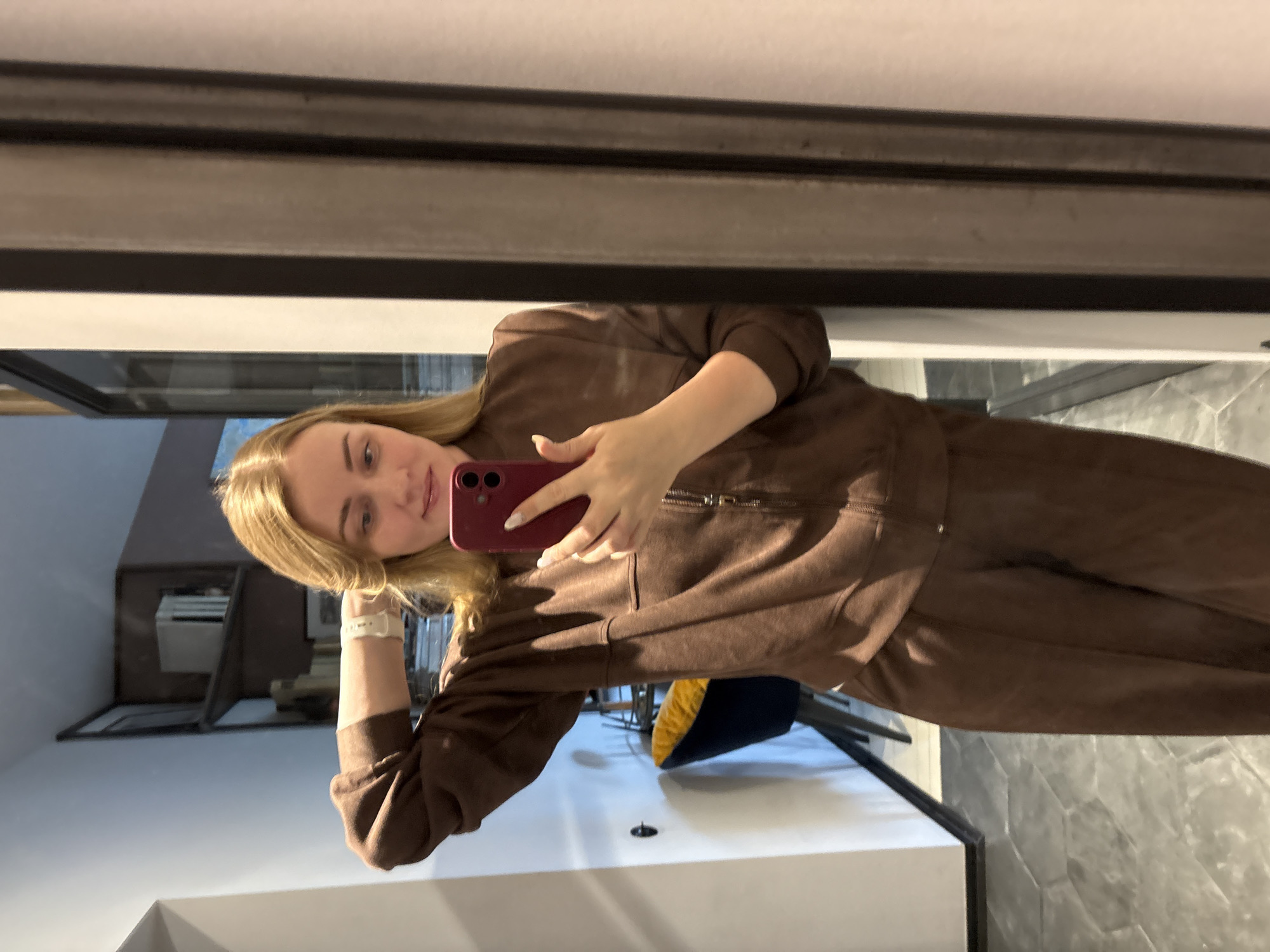 A young woman with blonde hair is taking a selfie in what appears to be a bathroom. She is wearing a brown sweatsuit and a white smartwatch. She is holding a red smartphone. The background features a modern bathroom design, including white walls, a grey tiled wall, and a glimpse of a blue and yellow chair. The image is reflected in a mirror with a black frame. It is impossible to determine the exact location in Saint Petersburg based on the provided image.