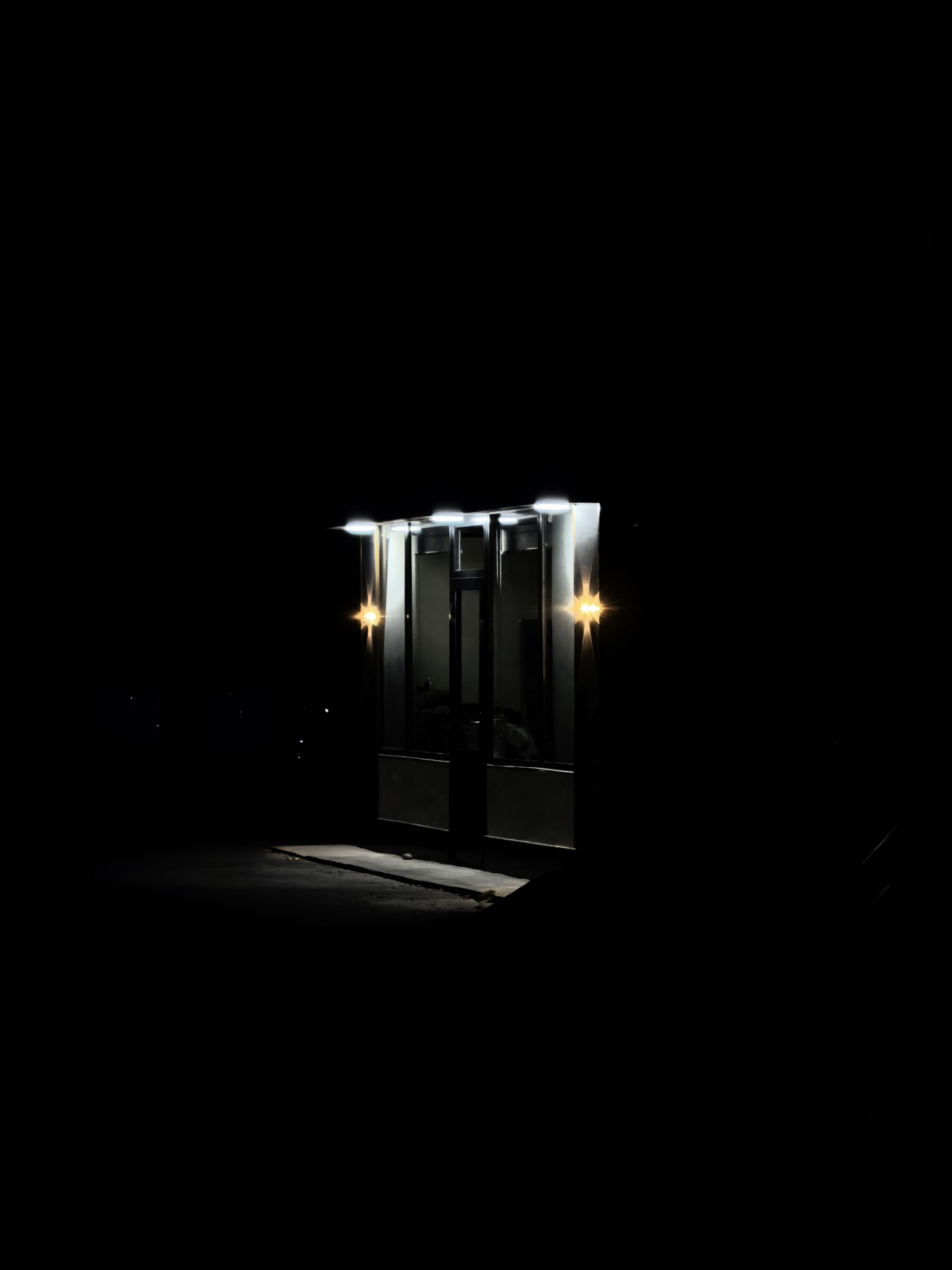A small, modern-style kiosk is illuminated at night in a dark outdoor setting. The kiosk has a prominent entrance with glass panels and a light fixture above, casting a bright glow. Two smaller, star-shaped lights flank the entrance, one on each side, adding to the illumination. The surrounding environment is predominantly black, suggesting a lack of ambient light, with faint, distant lights possibly indicating other structures or streetlights. The ground in front of the kiosk is visible, showing a paved or concrete surface with some unevenness. There are no individuals clearly discernible within or immediately around the kiosk, though faint shapes in the reflection of the glass might suggest a presence. The overall scene conveys a sense of isolation and nighttime commerce or service. The location is identified as Fergana City, Uzbekistan.