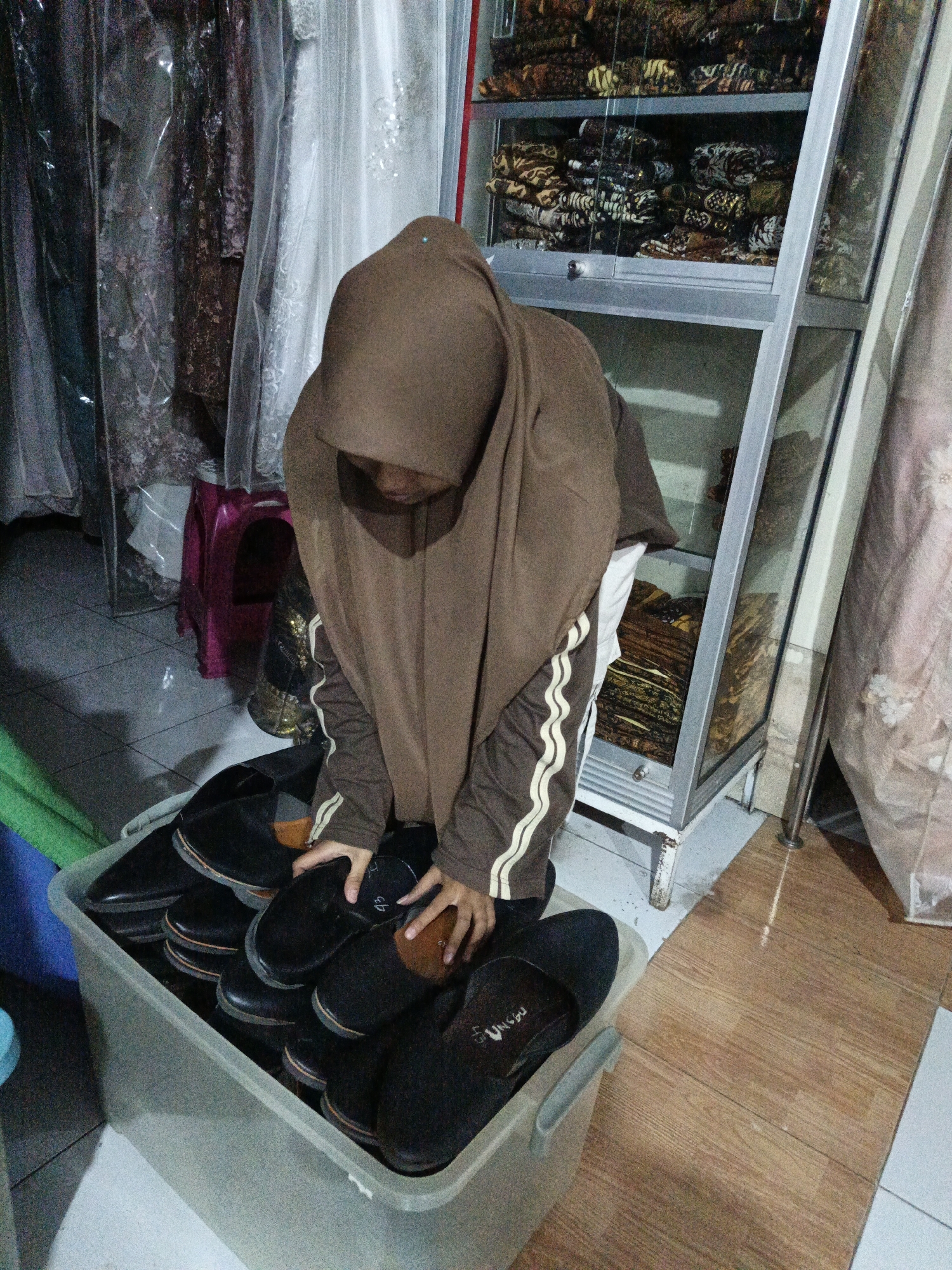 A young person wearing a brown hijab and a brown and white striped jacket is crouched down, reaching into a large, grey plastic container filled with black shoes. The container is overflowing with the shoes, which appear to be dress shoes or loafers. The scene takes place indoors, with racks of patterned fabrics visible in the background, suggesting a shop or a storage area for clothing. A pink stool is also visible to the left, partially obscured by plastic-wrapped garments. The flooring appears to be tile. The overall impression is a utilitarian space, possibly related to the sale or organization of footwear and textiles. The location is identified as Karangpakel, Indonesia.