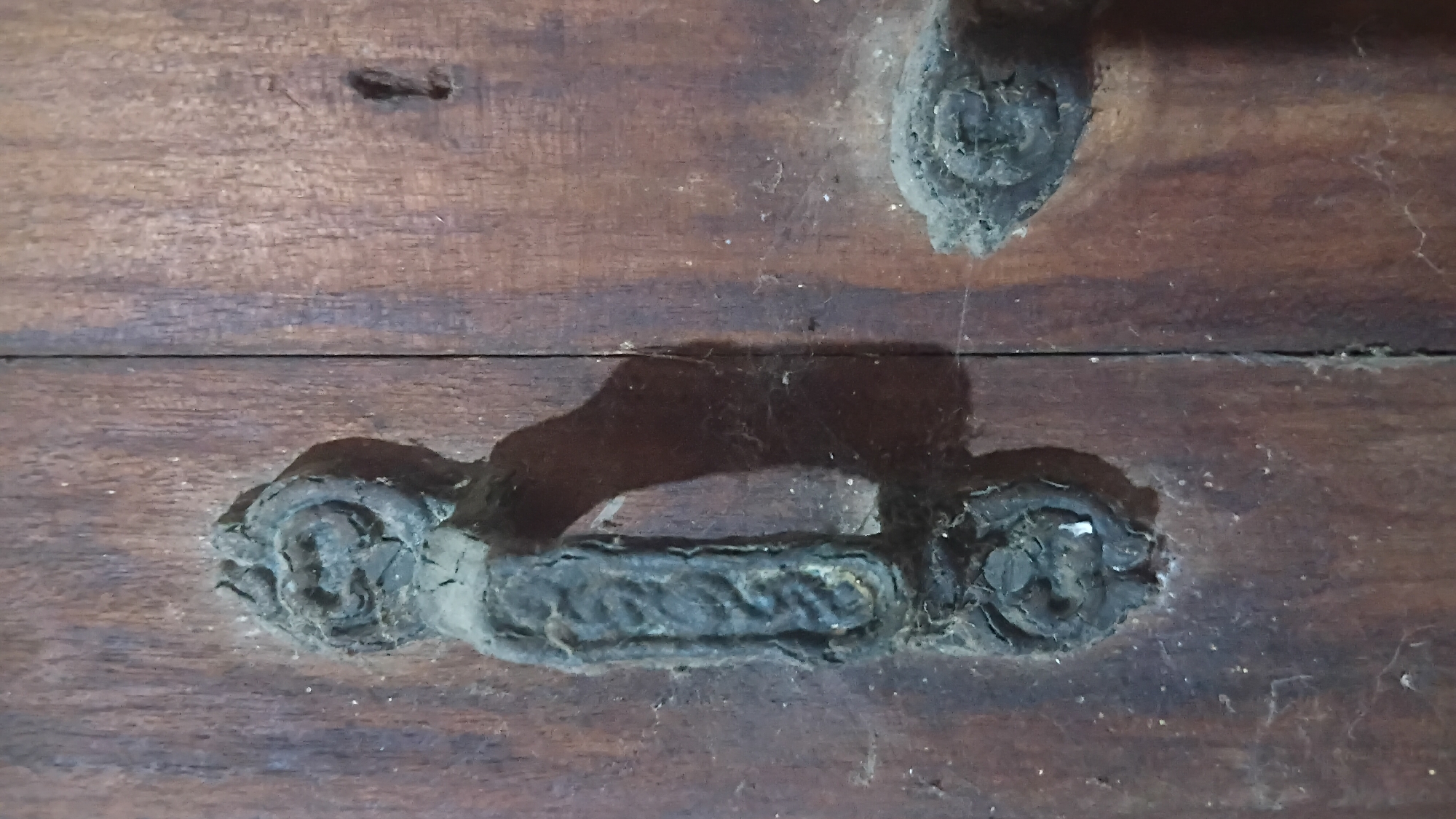 The image shows a close-up of an old, weathered wooden door with ornate, dark metal hardware. A vertical seam runs down the center of the door. On the left is a substantial door handle with a decorative, looped top and bottom section, and a textured, rectangular grip in between. The handle casts a shadow to the right. To the right of the handle, further down the door, is another piece of decorative metal hardware, possibly a keyhole cover or a decorative escutcheon, also with a circular, ornate design. A keyhole is visible to the right of this second piece of hardware. The wood grain of the door is prominent, and there are visible signs of age and wear, including dust and cobwebs clinging to the hardware and the door surface. The overall impression is one of age and a rustic aesthetic. The location is identified as Prabumulih, Indonesia.
