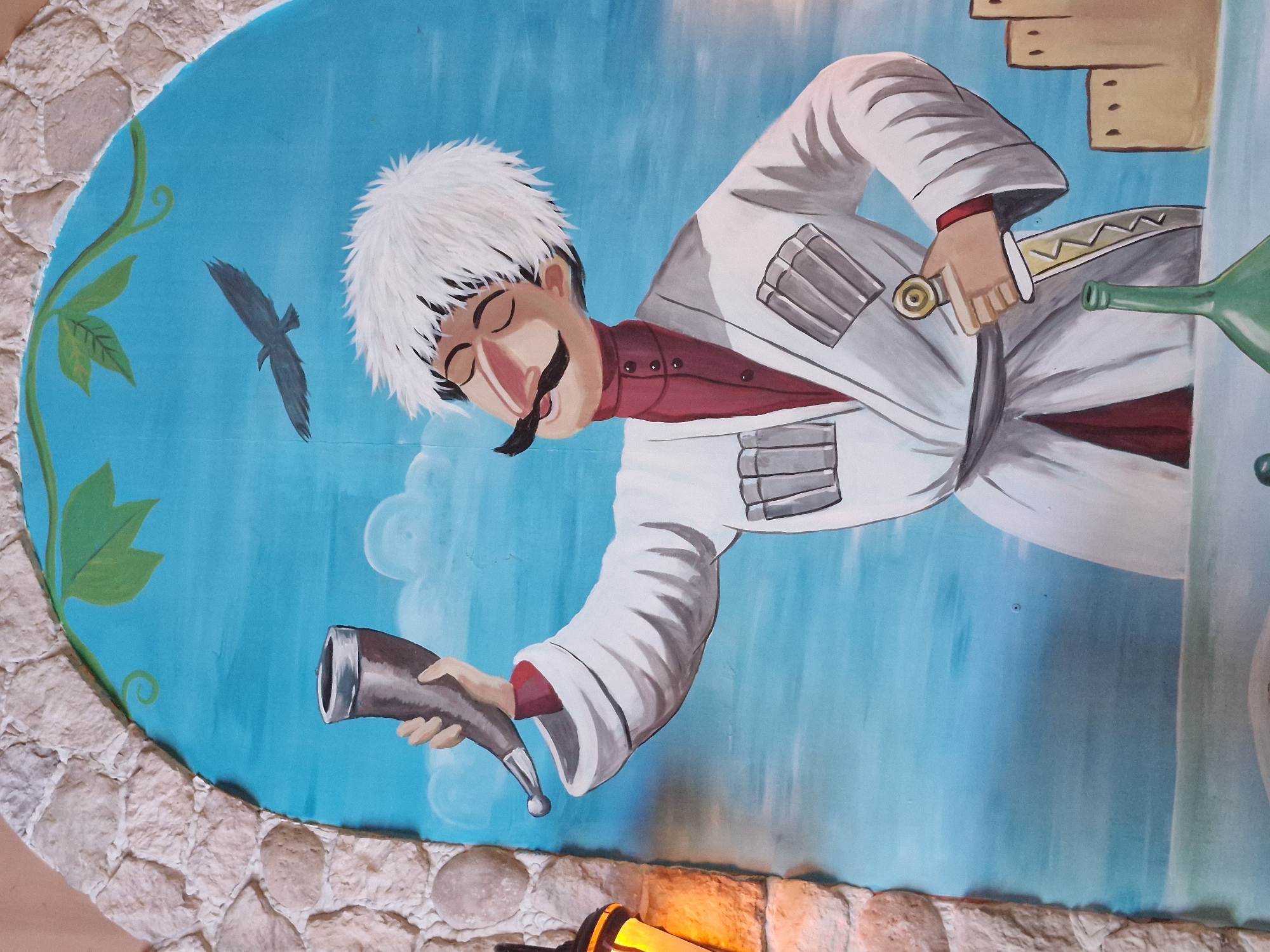 A mural depicts a cartoonish man in traditional attire, holding a drinking horn in his raised right hand. He wears a white robe over a dark red garment, a white fluffy hat, and has a prominent mustache. A dagger is held in his left hand. In the background, a bird flies across a bright blue sky with clouds. To the right, a stylized depiction of buildings can be seen. The mural is framed by a rough, stone-like archway. A lamp is visible on the left edge of the frame. The artwork appears to be located on an exterior wall, possibly in Kokshetau, Kazakhstan, given the context provided, but specific location details are not discernible from the image itself.