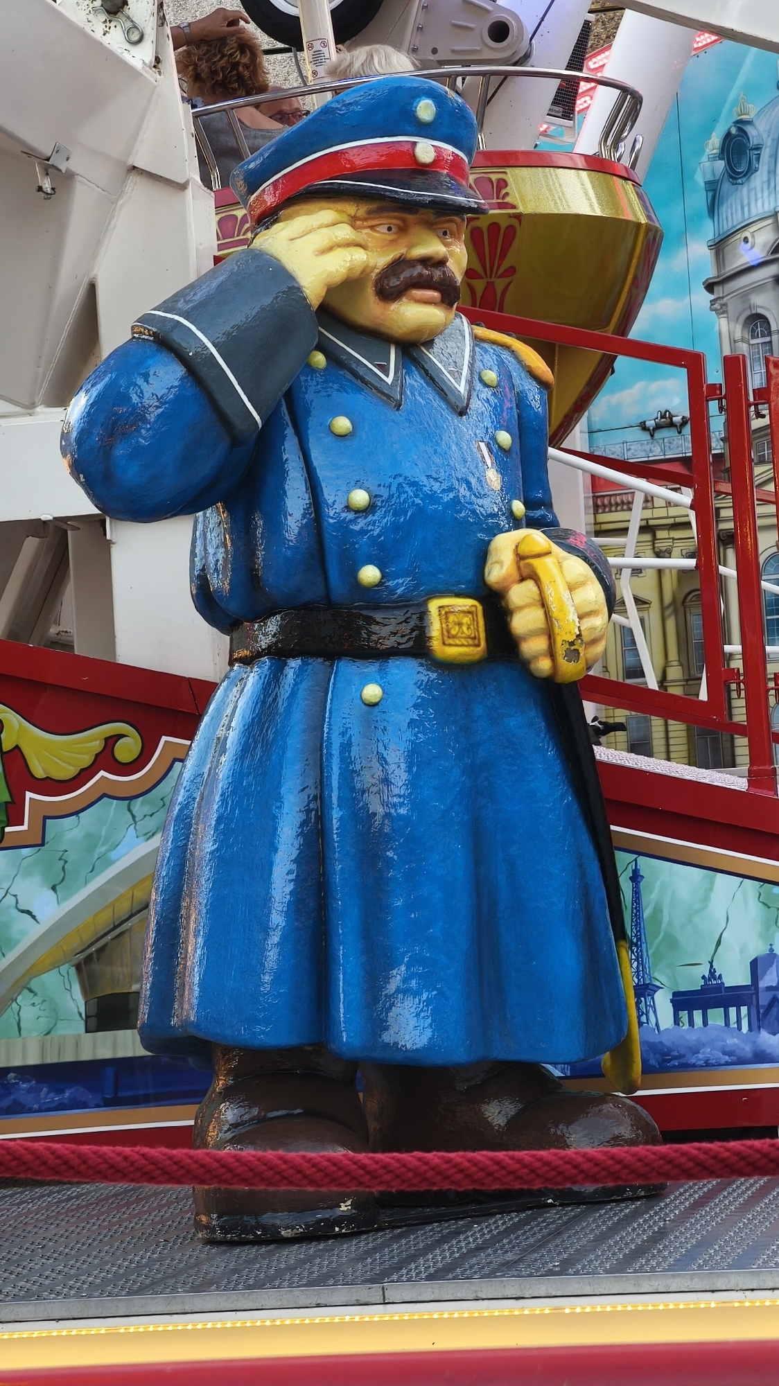 A life-sized, stationary, cartoonish sculpture of a man in a blue military-style uniform stands outdoors, with his right hand raised to his forehead in a salute. The sculpture has a yellow face with a dark mustache and a stern expression. He wears a blue cap with a red band and a gold emblem, a blue coat with gold buttons and epaulets, a black belt with a gold buckle, and dark brown boots. The backdrop consists of a brightly colored carnival ride with a red railing and painted scenery of buildings and an Eiffel Tower. The sculpture is positioned on a metallic platform, with a red rope barrier in the foreground. The lighting suggests it is daytime.