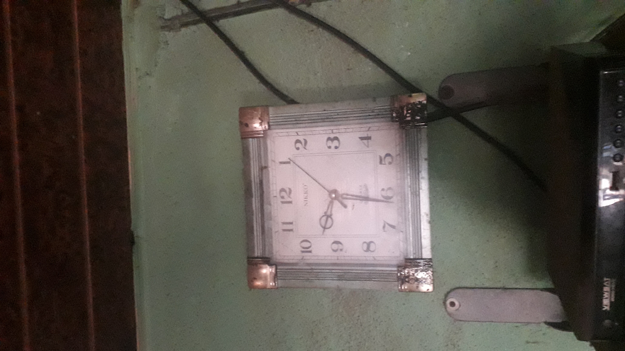 A square, analog wall clock with a white face and black numerals hangs on a textured, light green wall. The clock has gold-colored corner accents and a silver-toned frame with vertical fluting. The clock's hands indicate the time is approximately 7:08. Below the clock, on the wall, are two grey, elongated objects with circular indentations at their top ends, possibly speaker mounts or decorative elements. A dark, rectangular electronic device, labeled "VIEWSET" at the bottom, is positioned below these mounts. A dark cable runs diagonally across the right side of the frame. The environment appears to be indoors, possibly a room in Nkpor, Nigeria, given the context.