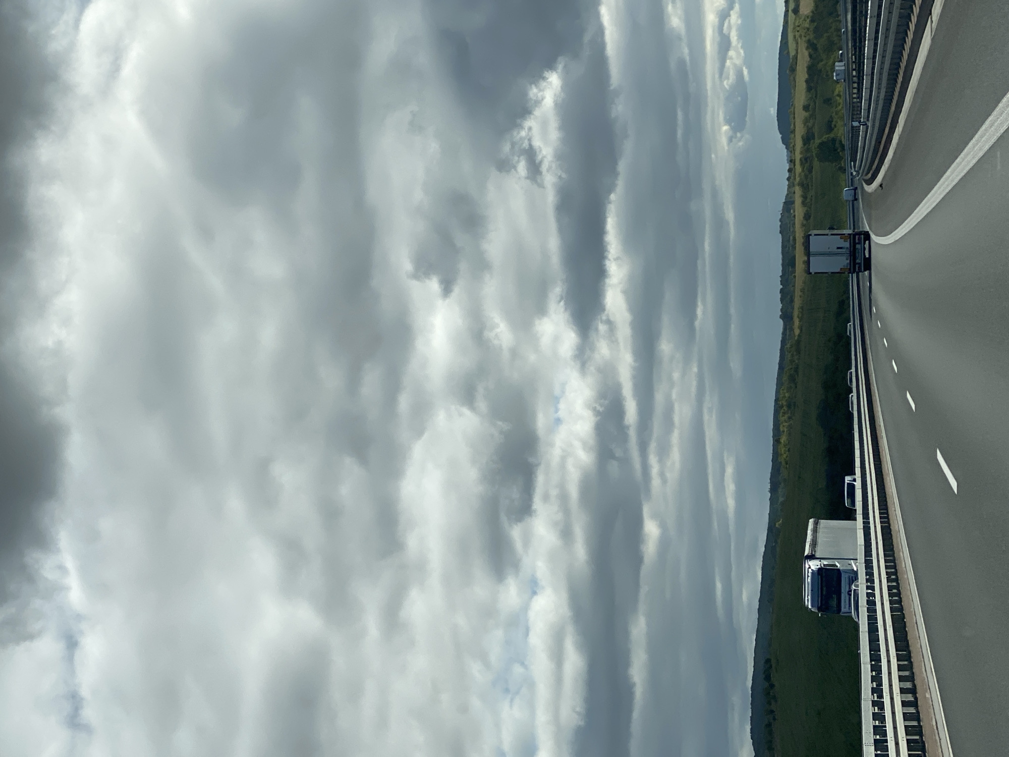 An overcast sky with dramatic, layered clouds stretches across the frame above a highway scene. In the foreground, the highway is empty except for lane markings. In the middle ground, a white semi-truck with a trailer is visible in the left lane, traveling away from the camera. Further down the highway, another white semi-truck is in the right lane, also traveling away from the camera. The surrounding landscape consists of rolling green hills and fields. The scene is likely a rural area, possibly near Heringen/Helme, Germany, given the architecture and landscape typical of the region.