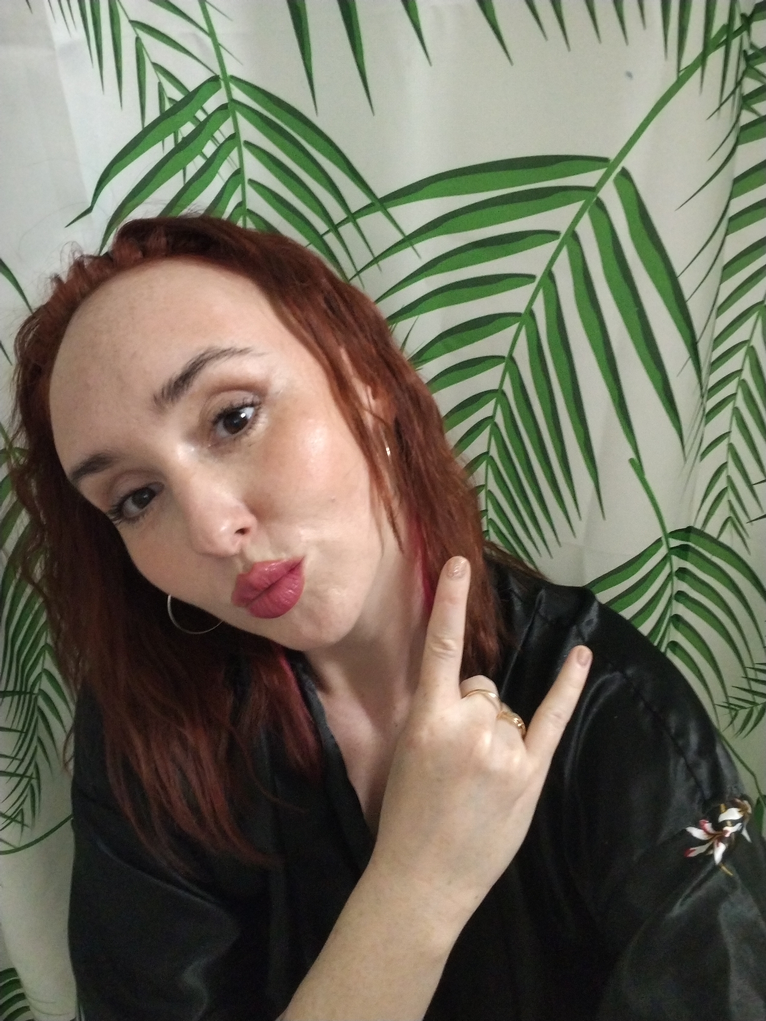 A fair-skinned woman with auburn hair is posing indoors against a shower curtain with a palm leaf pattern. She has freckles, hoop earrings, and is wearing a black silky robe or jacket. She is puckering her lips and making the "rock on" hand gesture with her right hand. A ring is visible on her right ring finger. The location appears to be a bathroom. The information about Izhevsk, Russia is not supported by the image.
