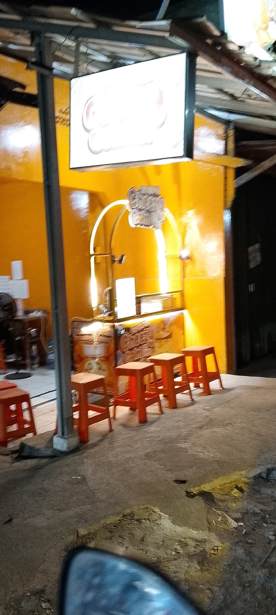 A low-angle, blurry, nighttime photograph captures a street-side food stall in Karangsatria, Indonesia. The stall, painted bright yellow, features a large, illuminated white sign with curved, stylized lettering that is unreadable due to the blur. Below the sign, a curved archway frames a service area with various unidentifiable objects and lights. Several orange plastic stools are lined up in front of the stall on a paved surface. The foreground shows a portion of a vehicle's interior, likely a car's dashboard and windshield, reflecting the scene. The overall impression is of a bustling, perhaps informal, nighttime food vendor.
