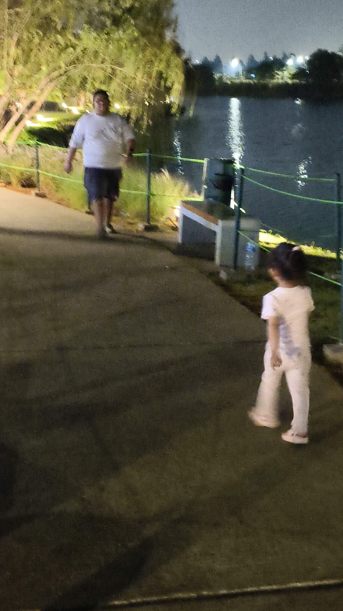 A full shot captures a walkway beside a body of water at night. In the foreground, a young girl in a white outfit walks away from the camera. Behind her, a man in a white t-shirt and dark shorts walks towards the camera. The walkway is made of dark asphalt. To the left, a tree with illuminated branches is visible. A fence with green ropes separates the walkway from the grassy area and the water. In the background, the water reflects light from distant lamps and the sky is dark. The scene appears to be a public park or recreational area. This image does not provide any information or clues that it is located in the city of West Jakarta, Indonesia.