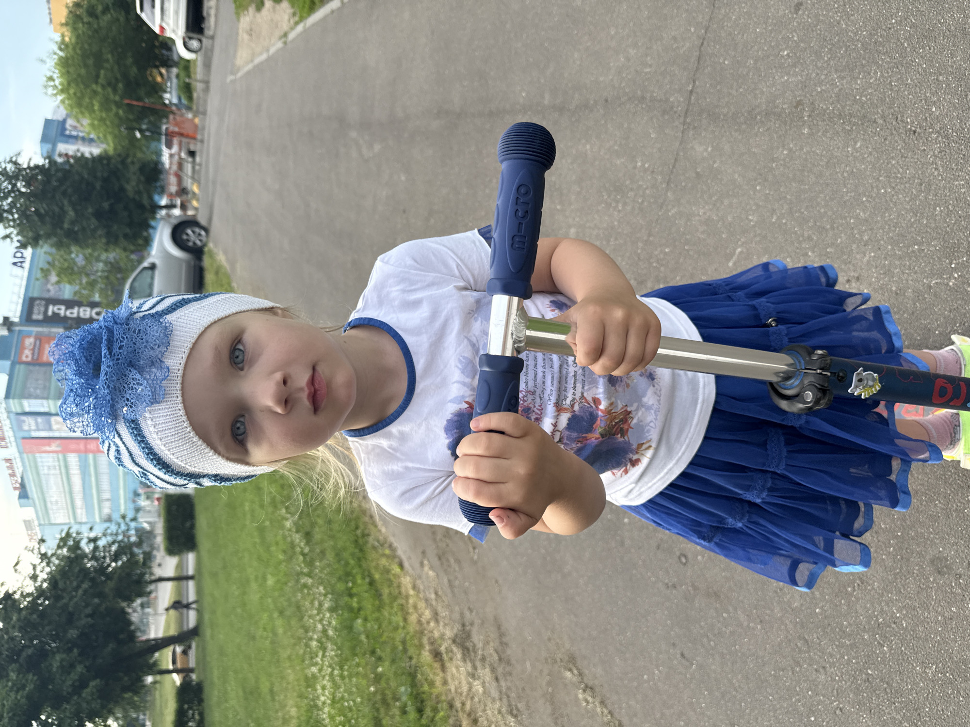A young girl with blonde hair, blue eyes, and a white t-shirt with a blue trim is standing on a paved path, holding the handlebars of a blue scooter. She is wearing a blue and white striped hat with a large blue flower embellishment and a full blue skirt. The background shows a grassy area, trees, and a large, modern building with blue and white accents. Cars are parked in the distance. The scene appears to be in an urban park or public space.